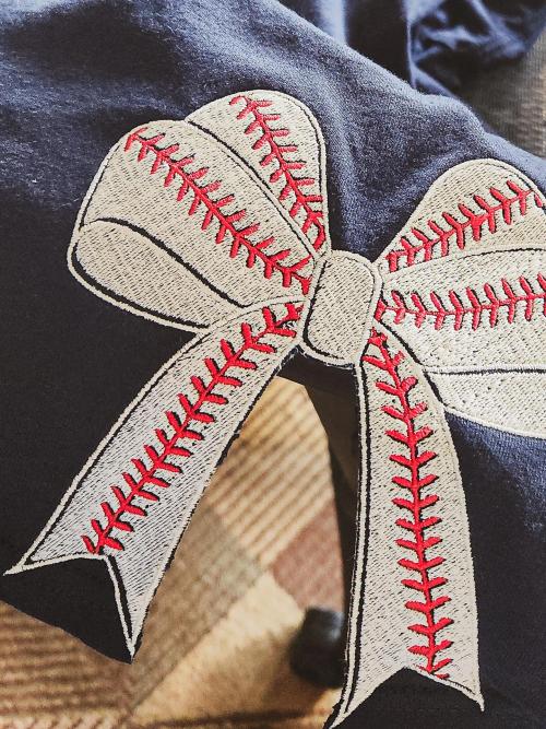 Large baseball-stitch-patterned bow appliqué with red stitching detail embroidered on a navy sweatshirt
