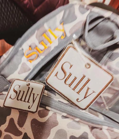 Orange script name "Sully" embroidered on a gray camo garment bag, with two matching leather-look "Sully" name tags hanging from the zipper pull