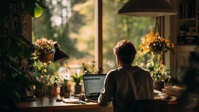 Person working thoughtfully at an organized desk with laptop displaying helpful AI interface elements, surrounded by natural lighting, plants, and personal touches in a comfortable workspace
