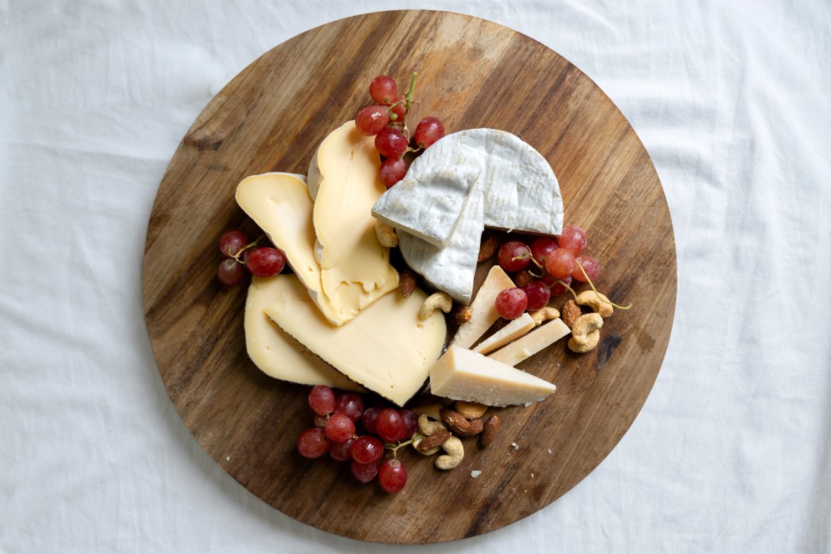 Overhead view of a round wooden board with assorted cheeses, red grapes and mixed nuts on white linen