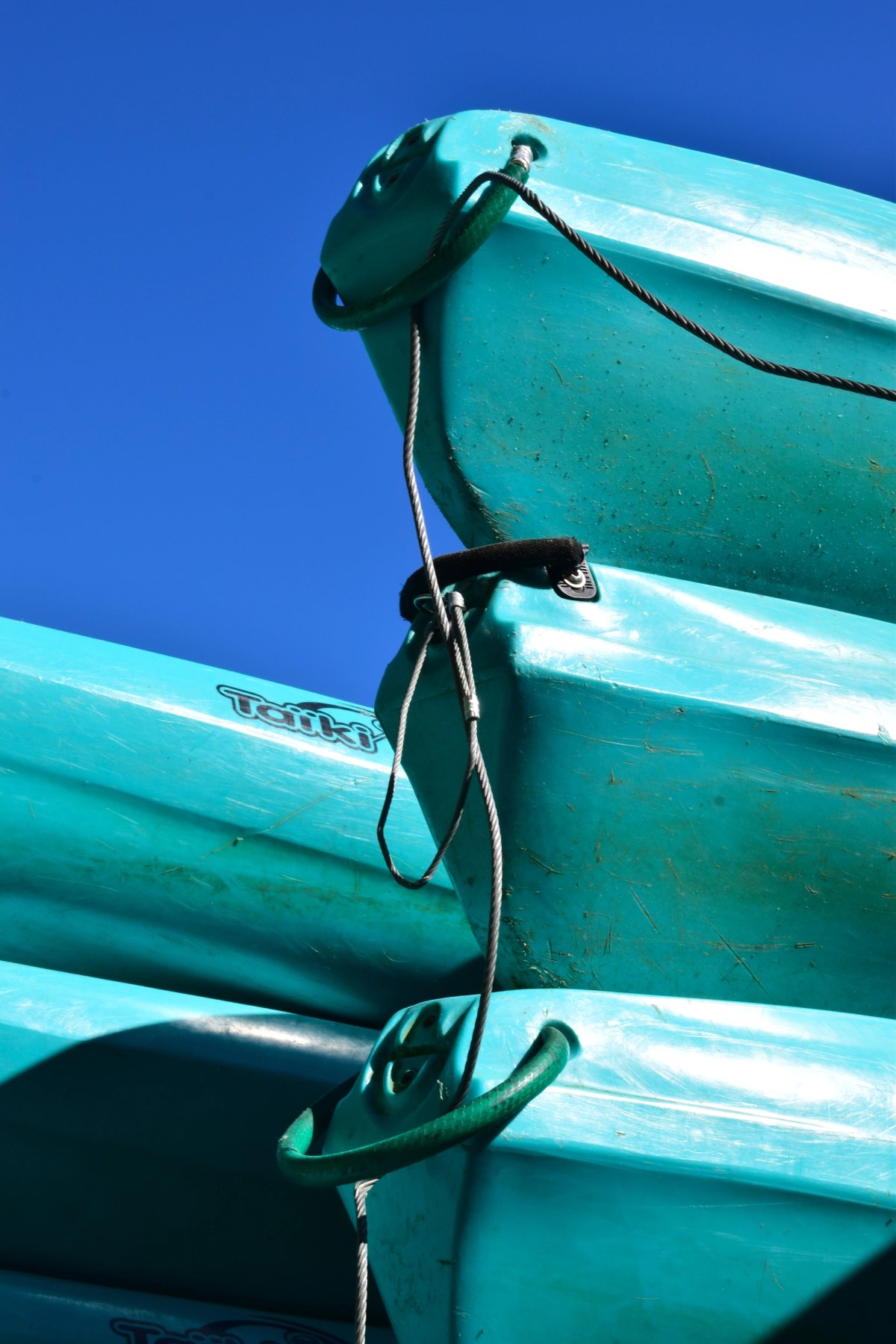 Stacked turquoise kayaks secured with rope and steel cable against a deep blue sky