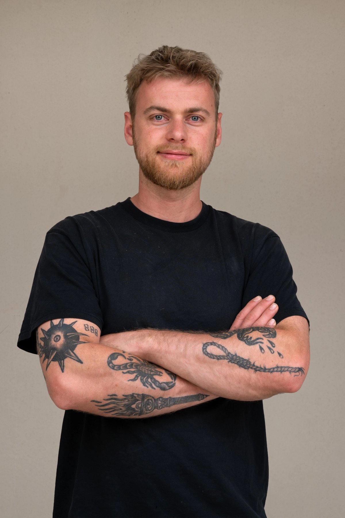 Studio portrait of a young man with a short beard and tattooed forearms, arms folded, on a warm grey backdrop