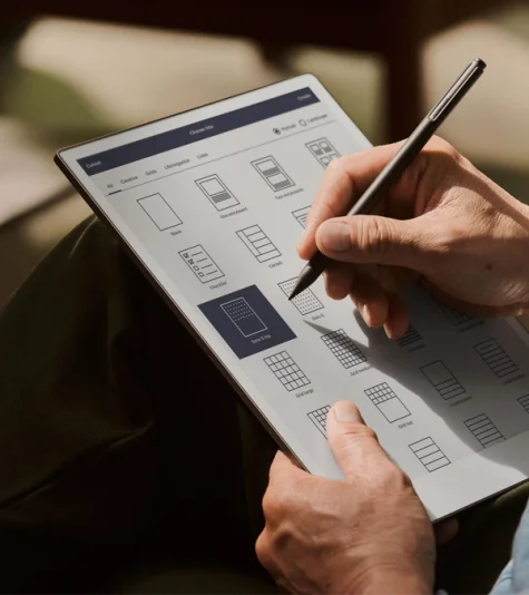 A close-up view of a person holding a reMarkable Paper Pro on their lap. Their finger is poised over the screen, which displays a grid of different note-taking templates, including a weekly planner and a meeting notes layout.
