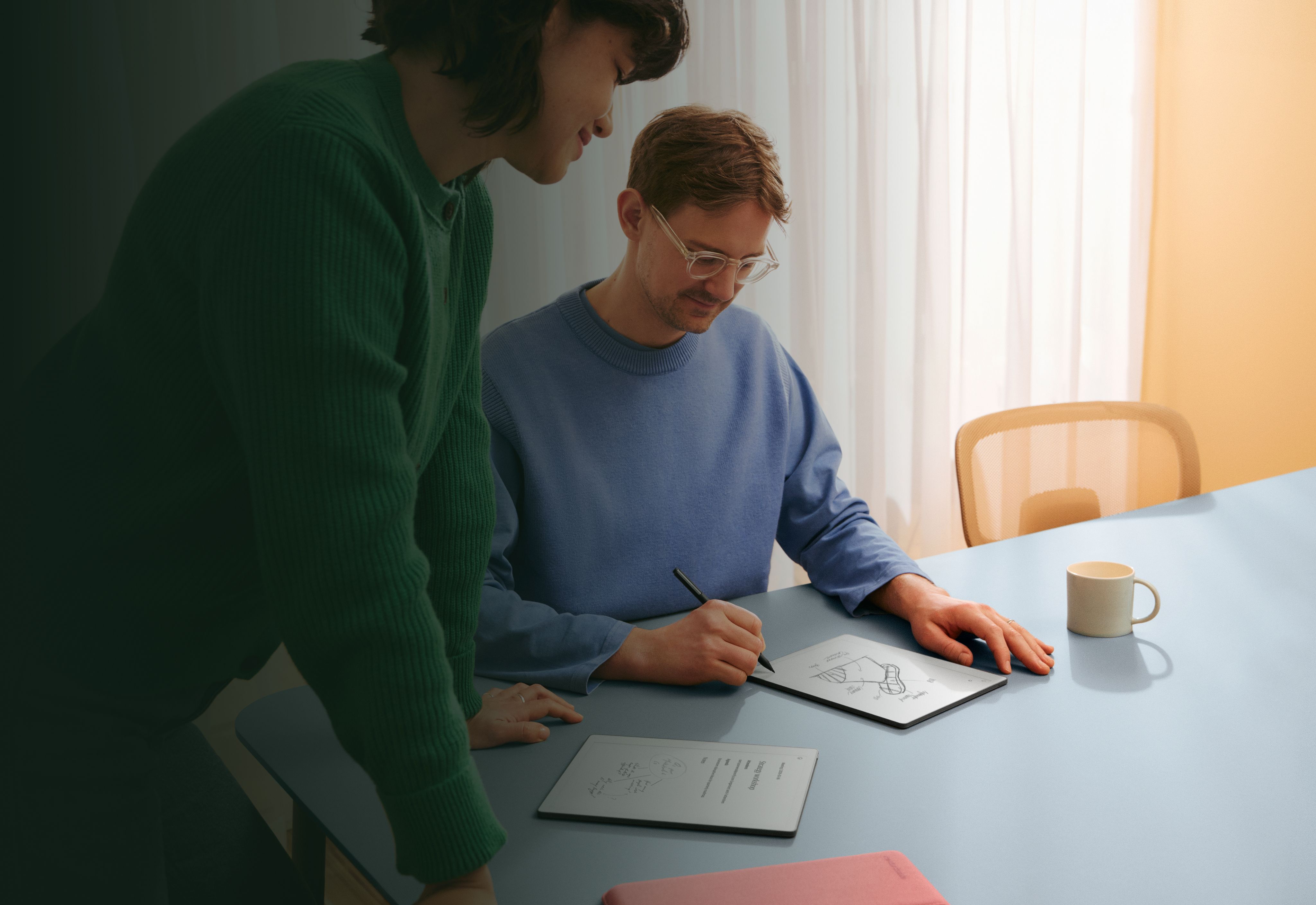 A man at a table writing on a reMarkable Paper tablet, in discussion with a woman looking at his notes