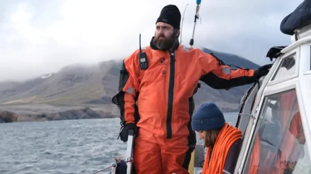 A man in an orange suit on a boat near a mountain.