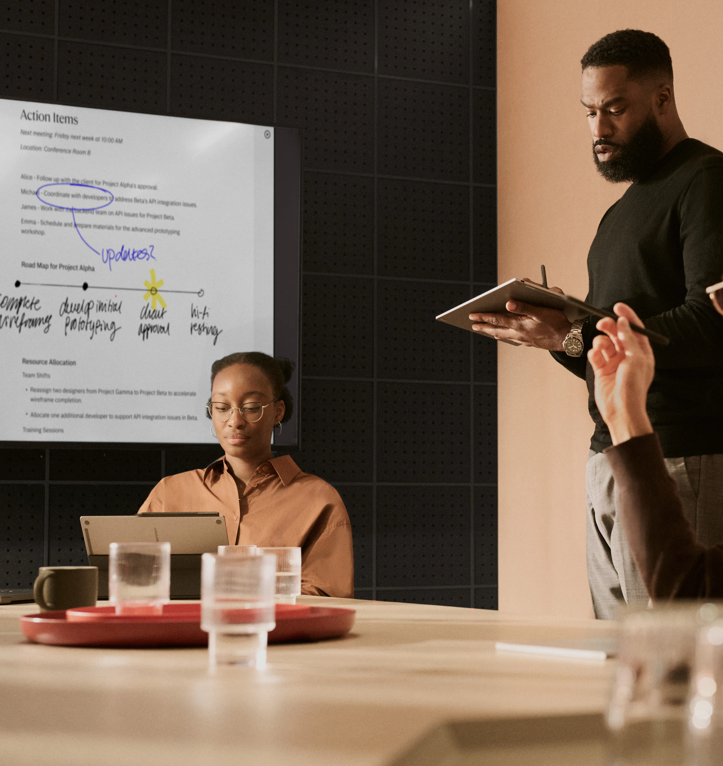 A professional team collaborating in a meeting room, featuring a large wall display sharing live annotated reMarkable notes with action items, while a man in the foreground takes notes on a reMarkable paper tablet.