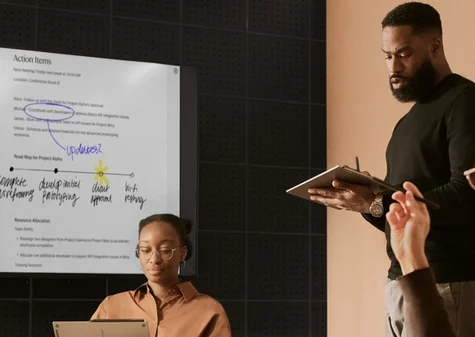 A professional team collaborating in a meeting room, featuring a large wall display sharing live annotated reMarkable notes with action items, while a man in the foreground takes notes on a reMarkable paper tablet.