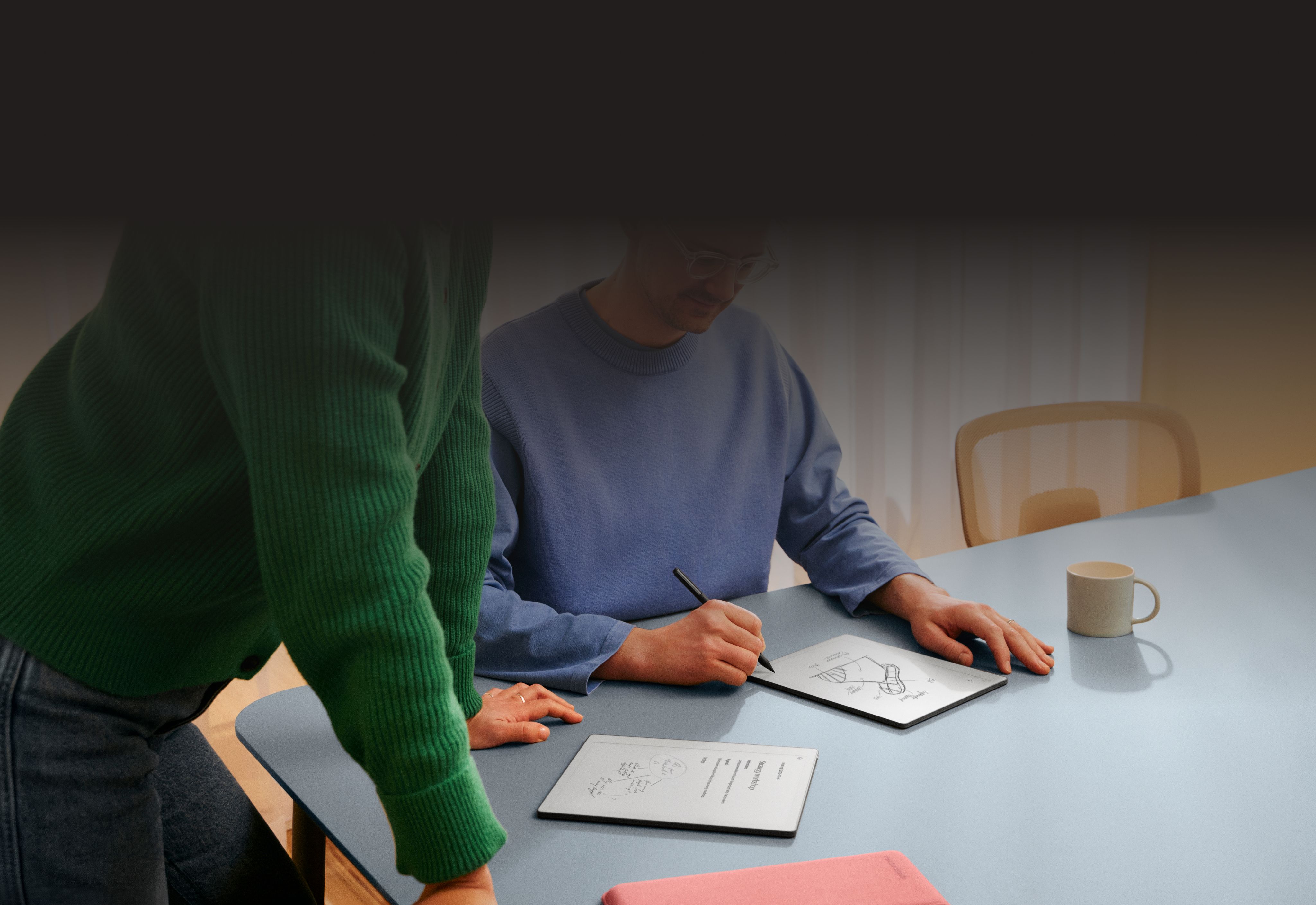 A man at a table writing on a reMarkable Paper tablet, in discussion with a woman looking at his notes