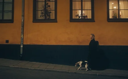 Anna walking her dog by buildings with lit windows in the evening.