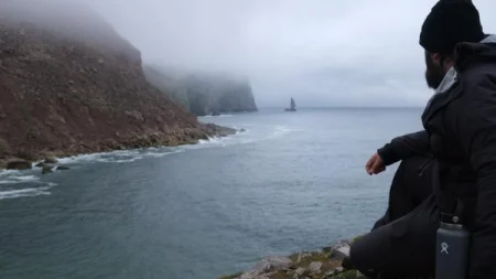 A man sits by the sea and looks out at a distant sailboat.