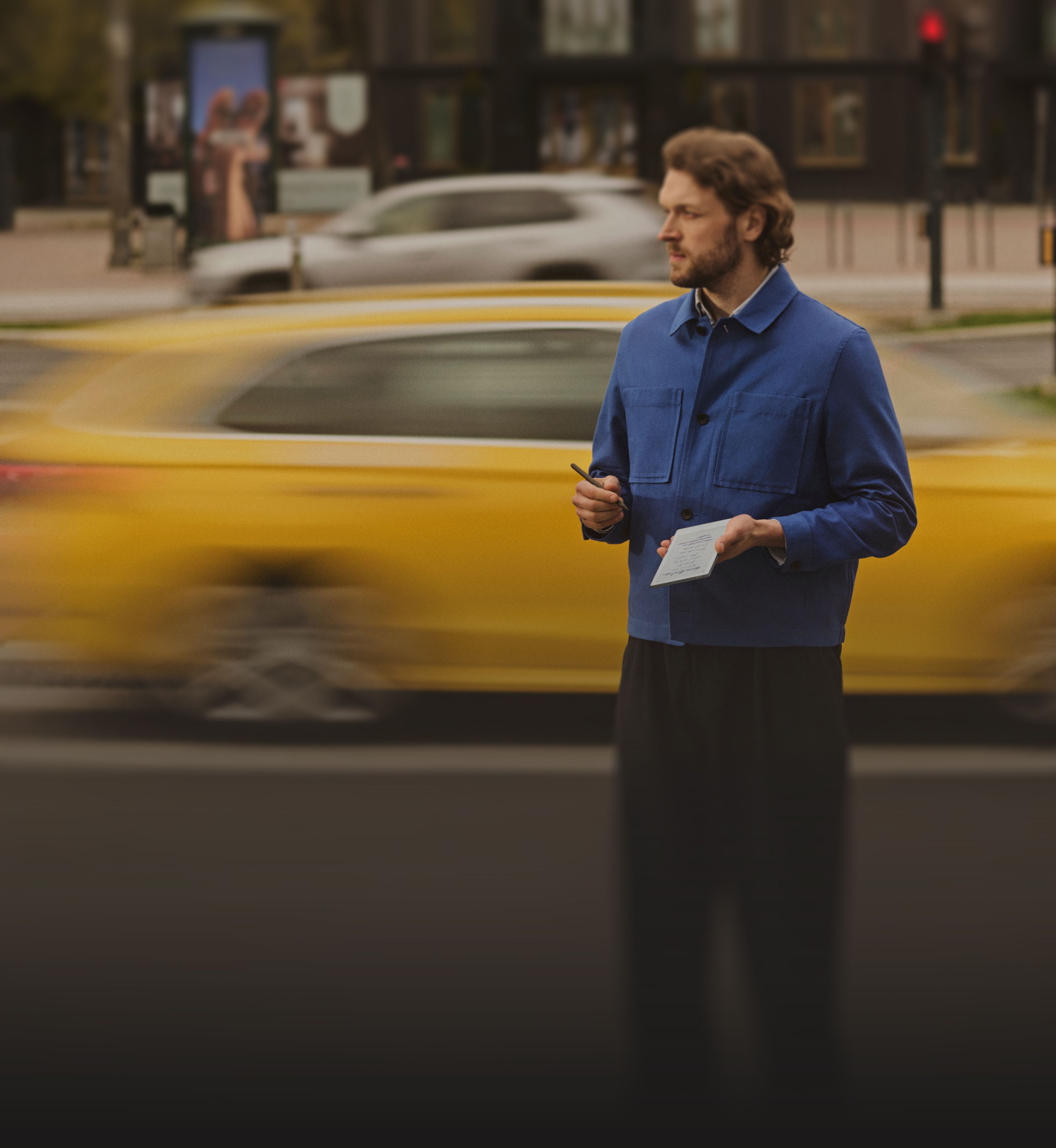 A man standing on a street corner holding a reMarkable Paper Pro Move paper tablet, with traffic moving behind him.