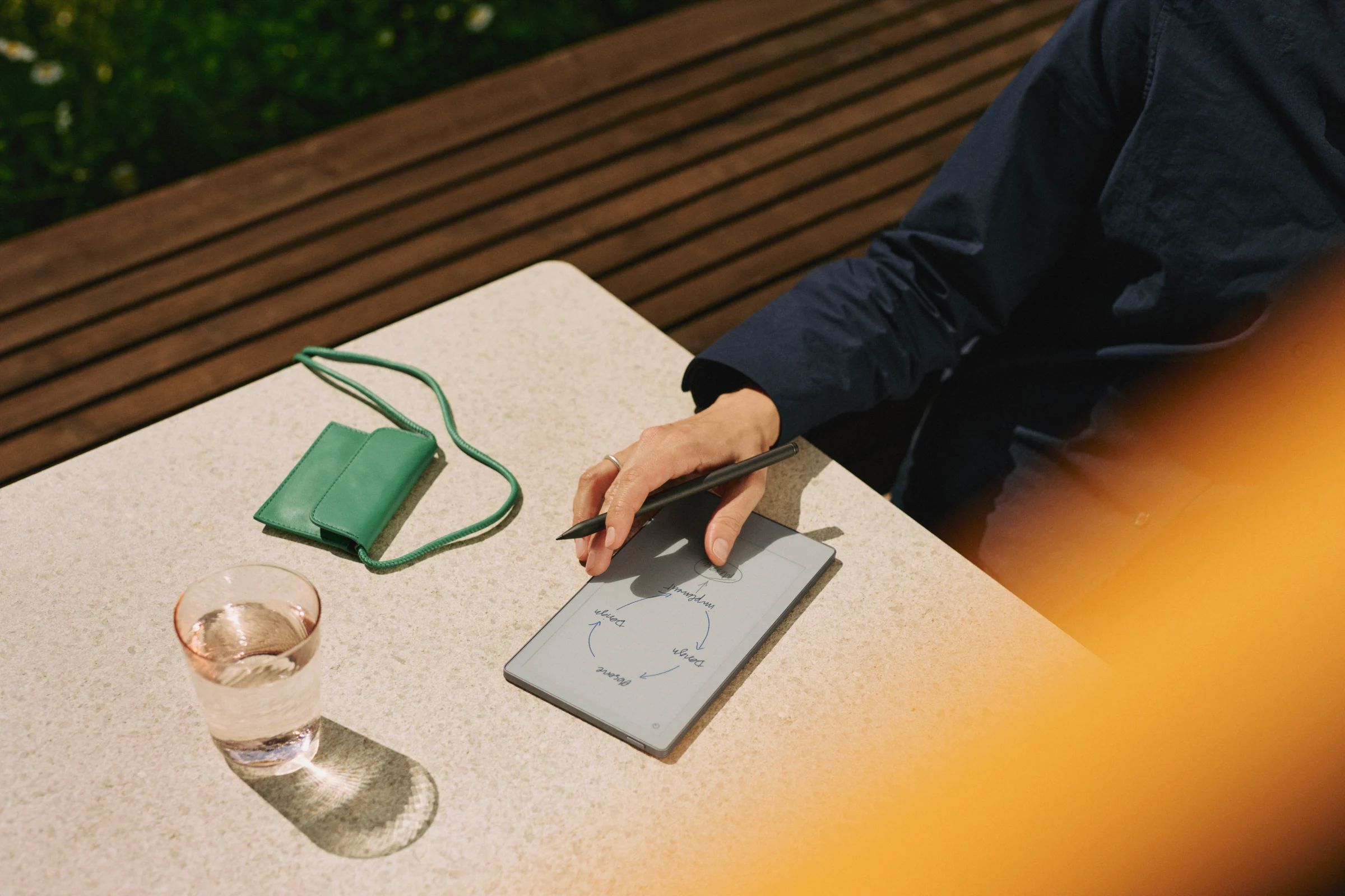 Top-down view of a hand drawing a flowchart diagram with a Marker Plus on a reMarkable Paper Pro Move set on an outdoor stone table alongside a glass of water and a green leather accessory pouch with a strap.