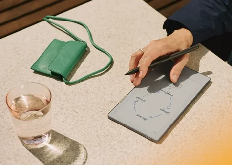 Top-down view of a hand drawing a flowchart diagram with a Marker Plus on a reMarkable Paper Pro Move set on an outdoor stone table alongside a glass of water and a green leather accessory pouch with a strap.