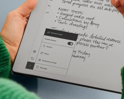 Close-up view of hands holding an reMarkable paper tablet with handwritten meeting notes, highlighting an open software overlay menu with options like Convert to text, Enable shapes toggle, Search, Share, and Notebook settings.