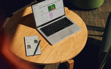 Top-down view of a round wooden table with a reMarkable paper tablet and Marker Plus next to an open laptop, showing file synchronization across both device screens.