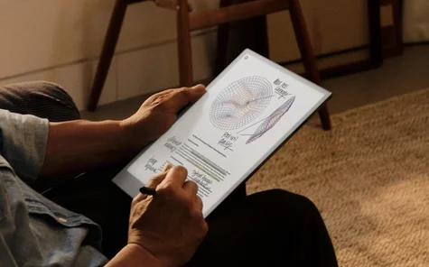 A person seated in warm indoor lighting, holding a reMarkable paper tablet displaying an annotated document with colored 3D wireframe mesh diagrams and handwritten notes, using a Marker Plus to write on the screen.