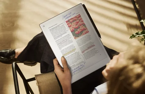 Over-the-shoulder view of a person seated and holding a reMarkable paper tablet displaying a formatted report with color photographs of flowers, handwritten blue annotations, and highlighted text.