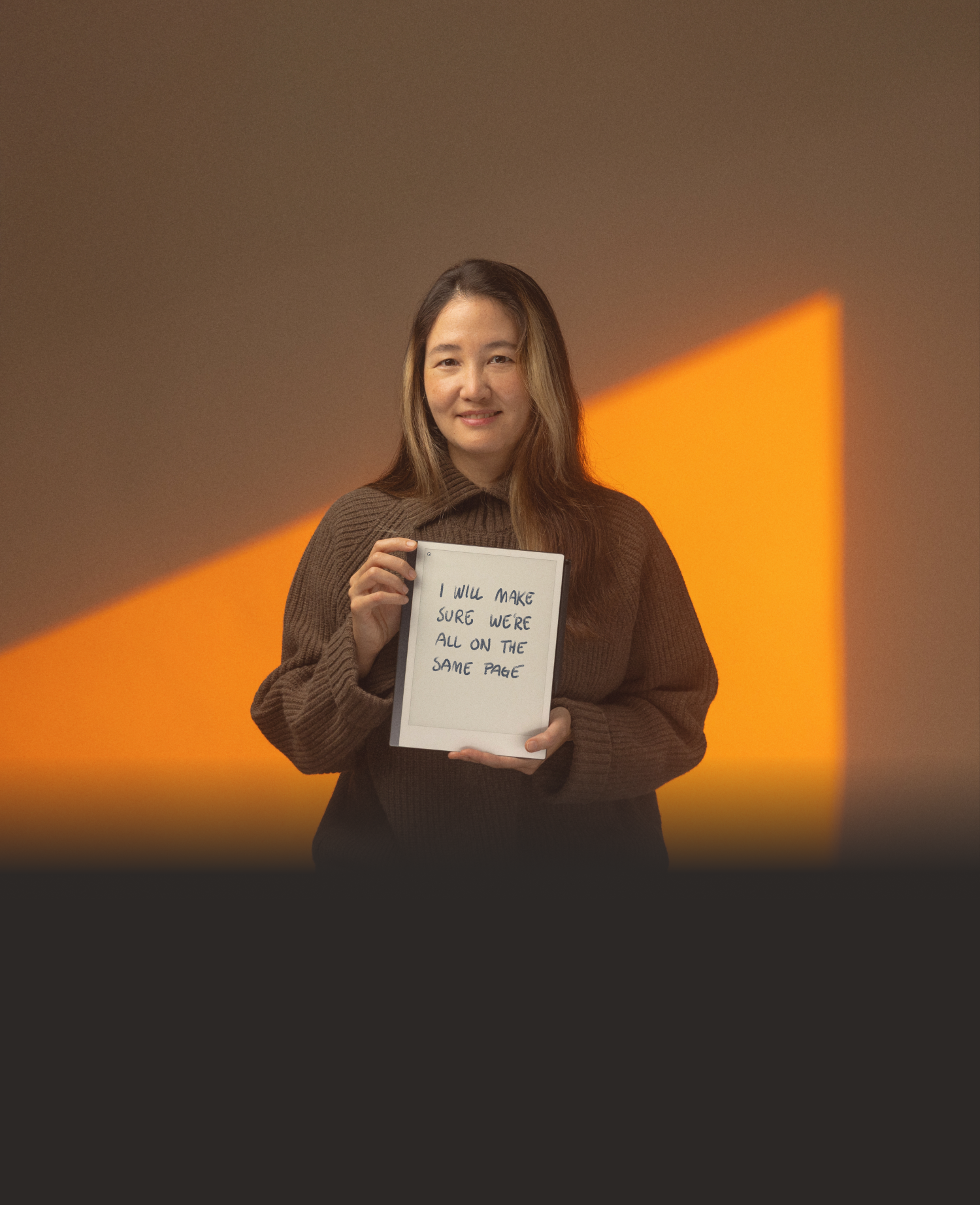 A woman holds a reMarkable paper tablet with the text "I will make sure we're all on the same page".
