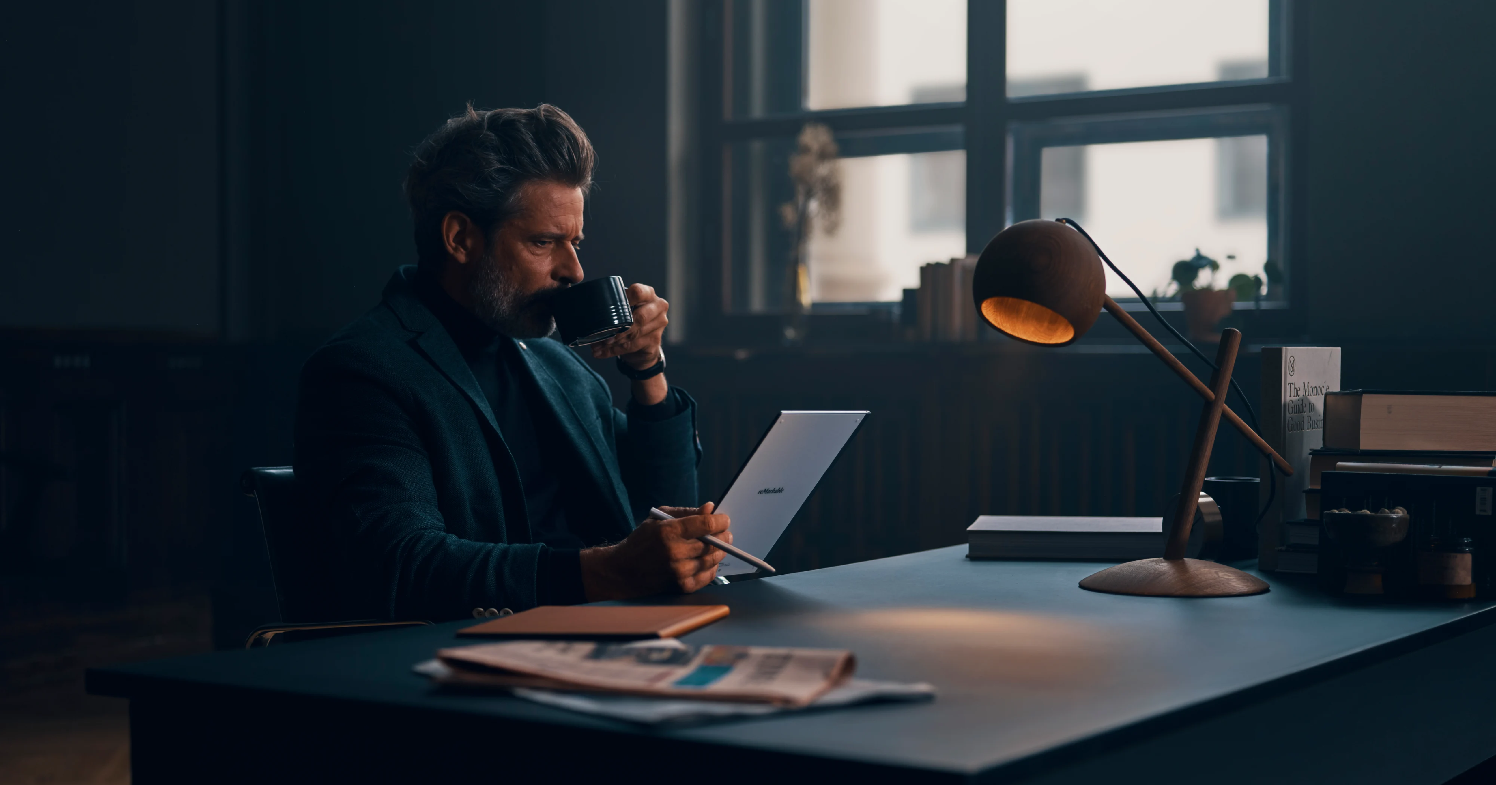 Man drinking coffee with reMarkable paper tablet. Newsprint and lamp on desk in moody office.