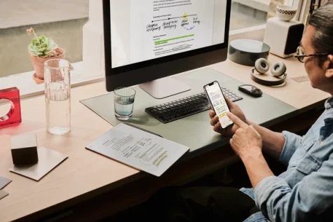 A man looking at a phone next to a computer displaying notes and a reMarkable paper tablet.