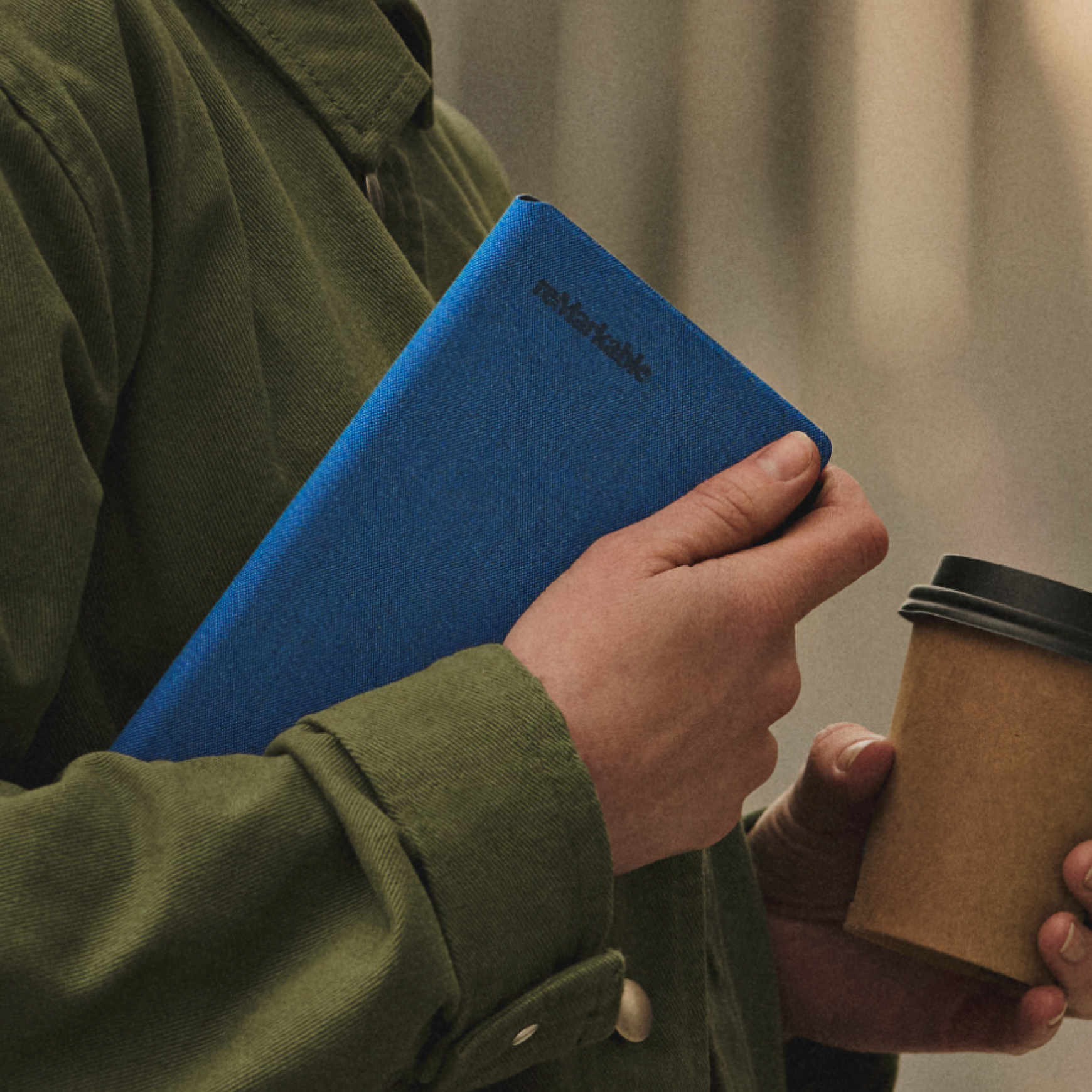 A woman outside with a green coat, holding a reMarkable Paper Pro Move in a cobalt blue Book Folio in one hand, and a coffee cup in the other.