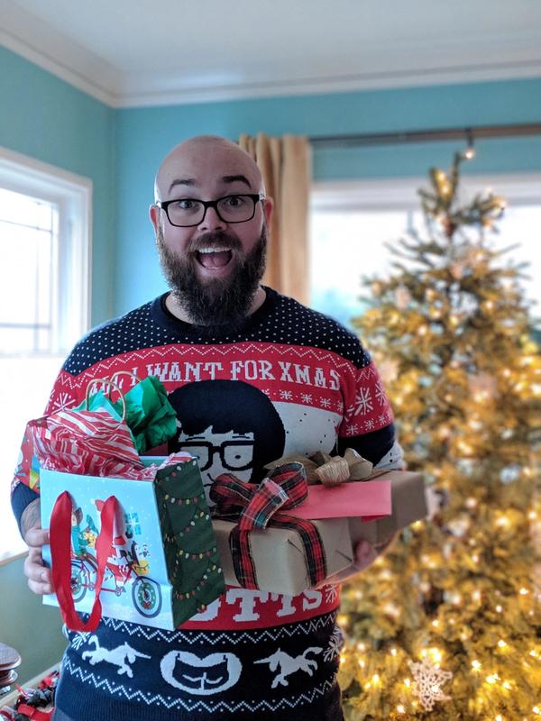 Jason standing in front of a Christmas tree holding several presents and smiling