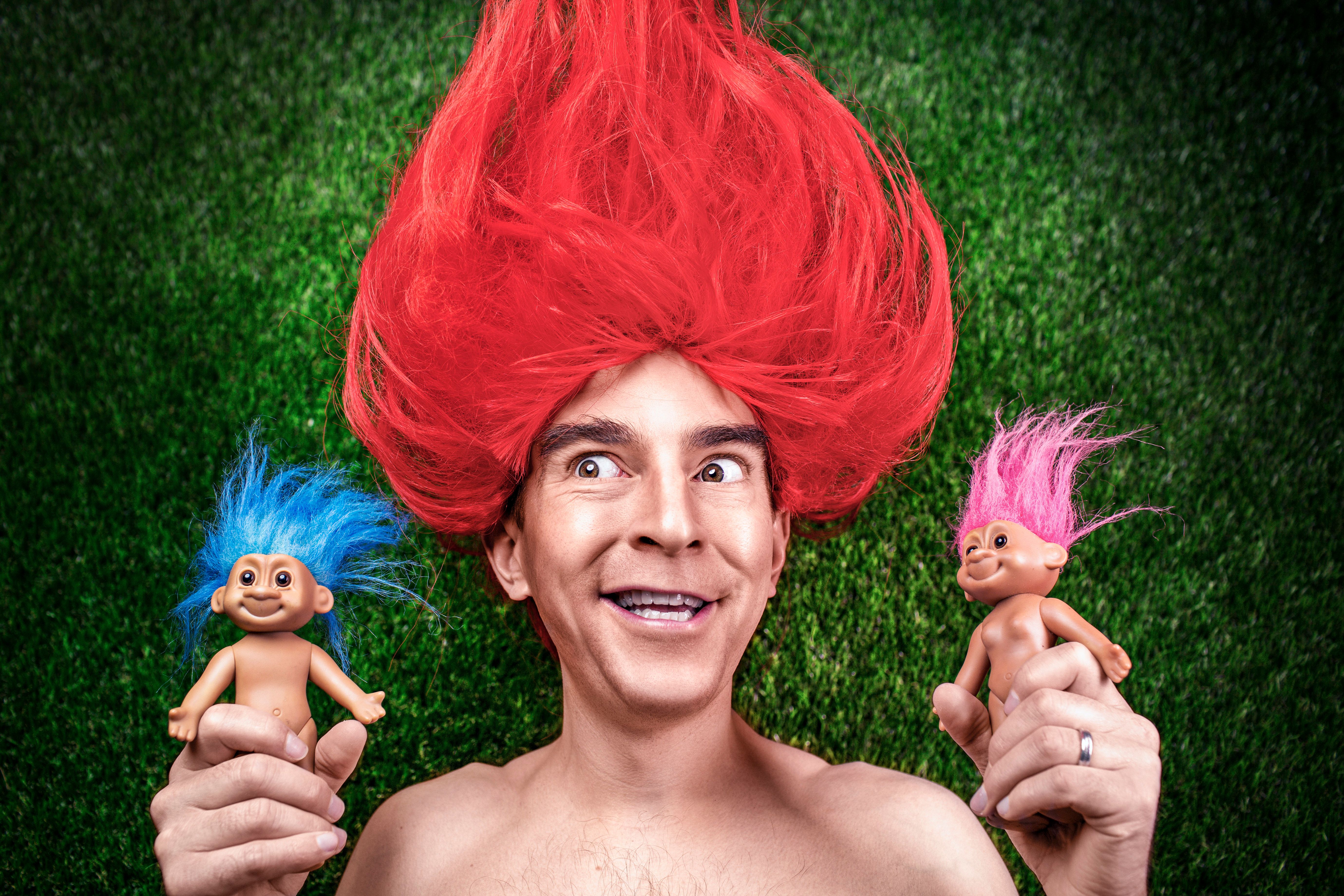 Man with red troll hair holding two troll dolls.