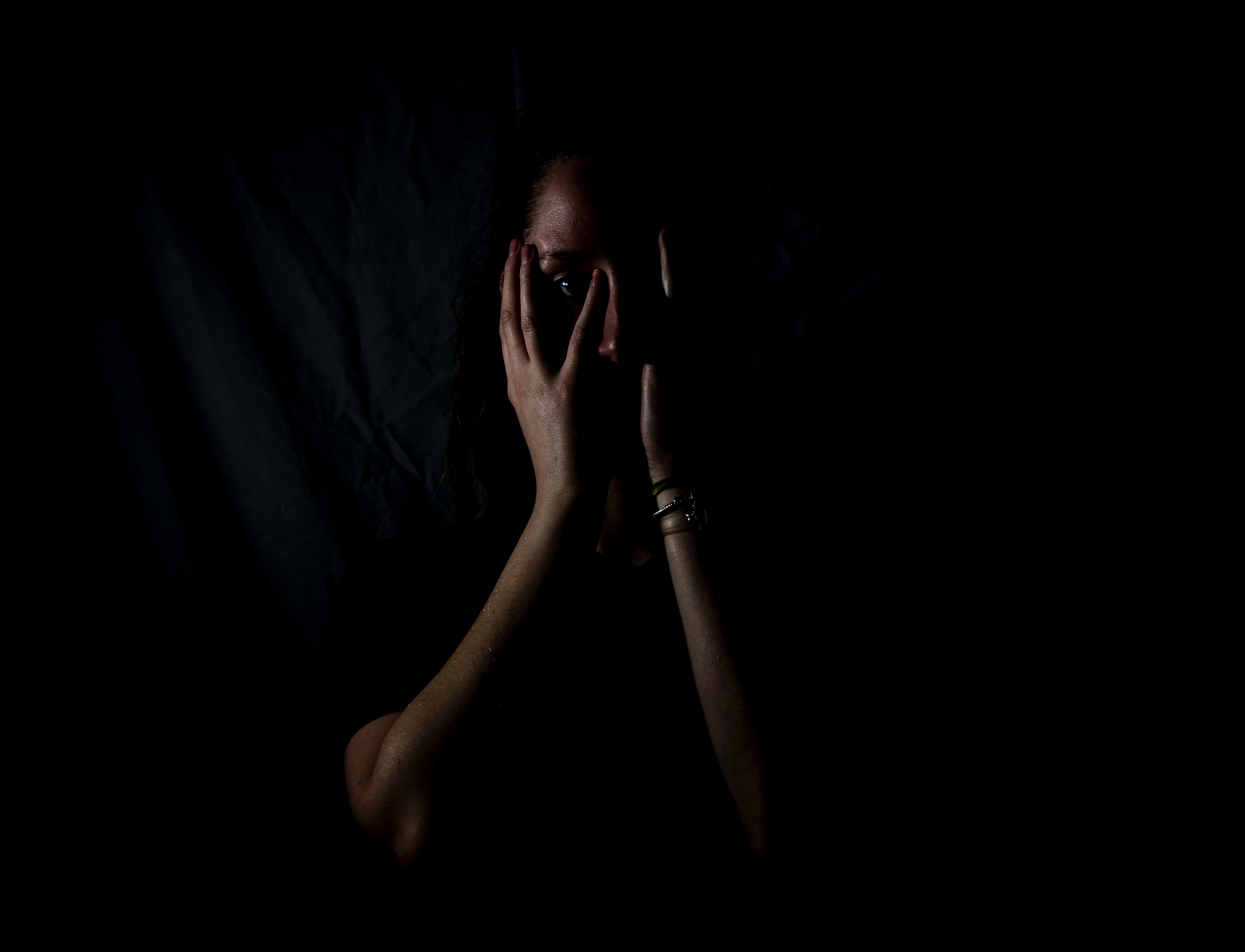 Person covering their face and standing in the dark, looking nervous.