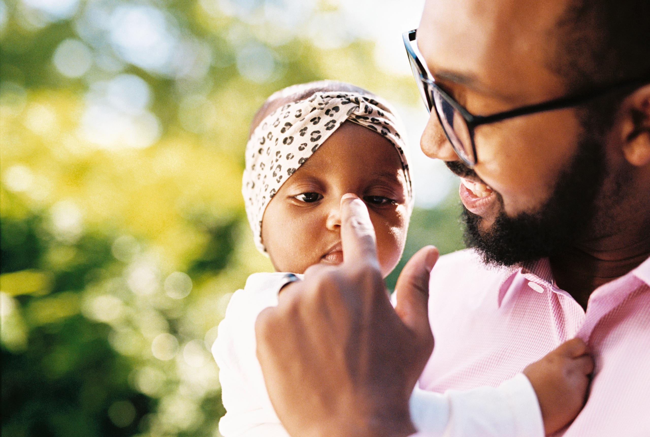A father holding a baby in his arms and pointing at the baby's nose.
