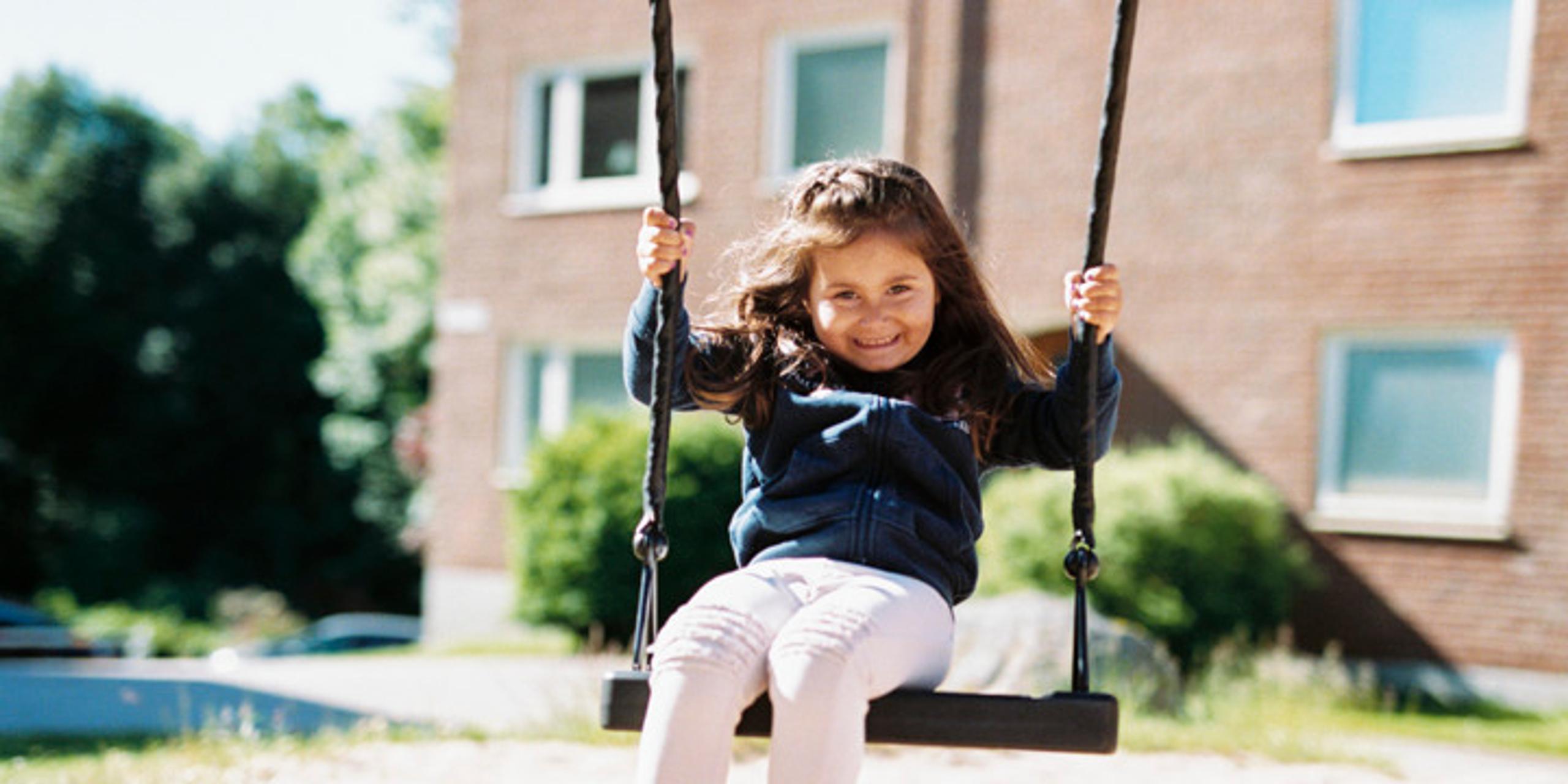 A girl on a swing at a playground in a residential area, smiling and looking into the camera.