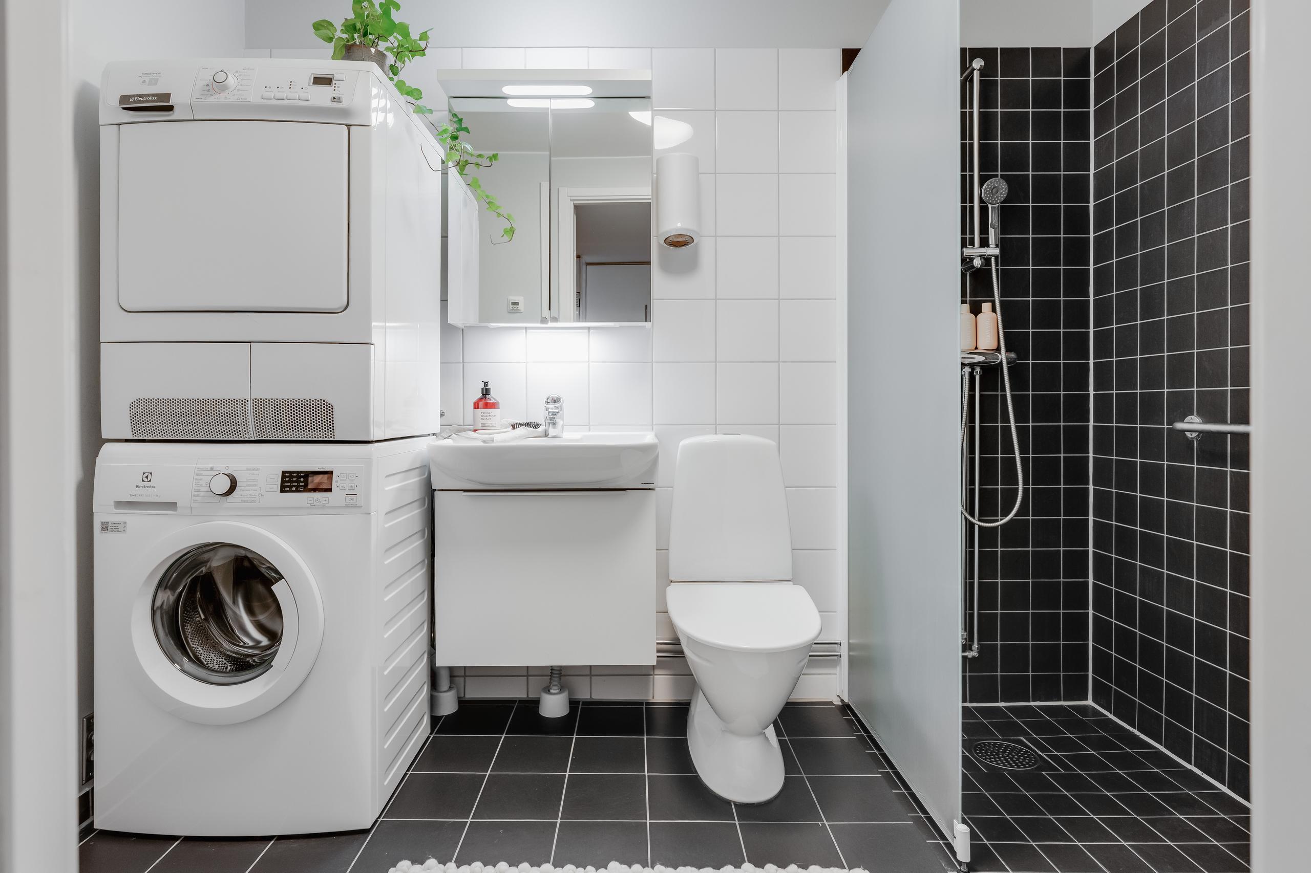 Badrum i vitt och svart kakel. Tvättmaskin, torktumlare, handfat, wc och dusch.