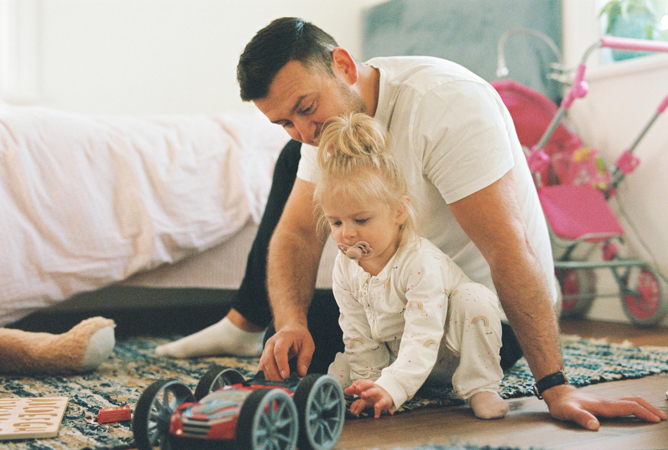 A man and a small girl sit on the floor of a child's room, playing with a toy car.