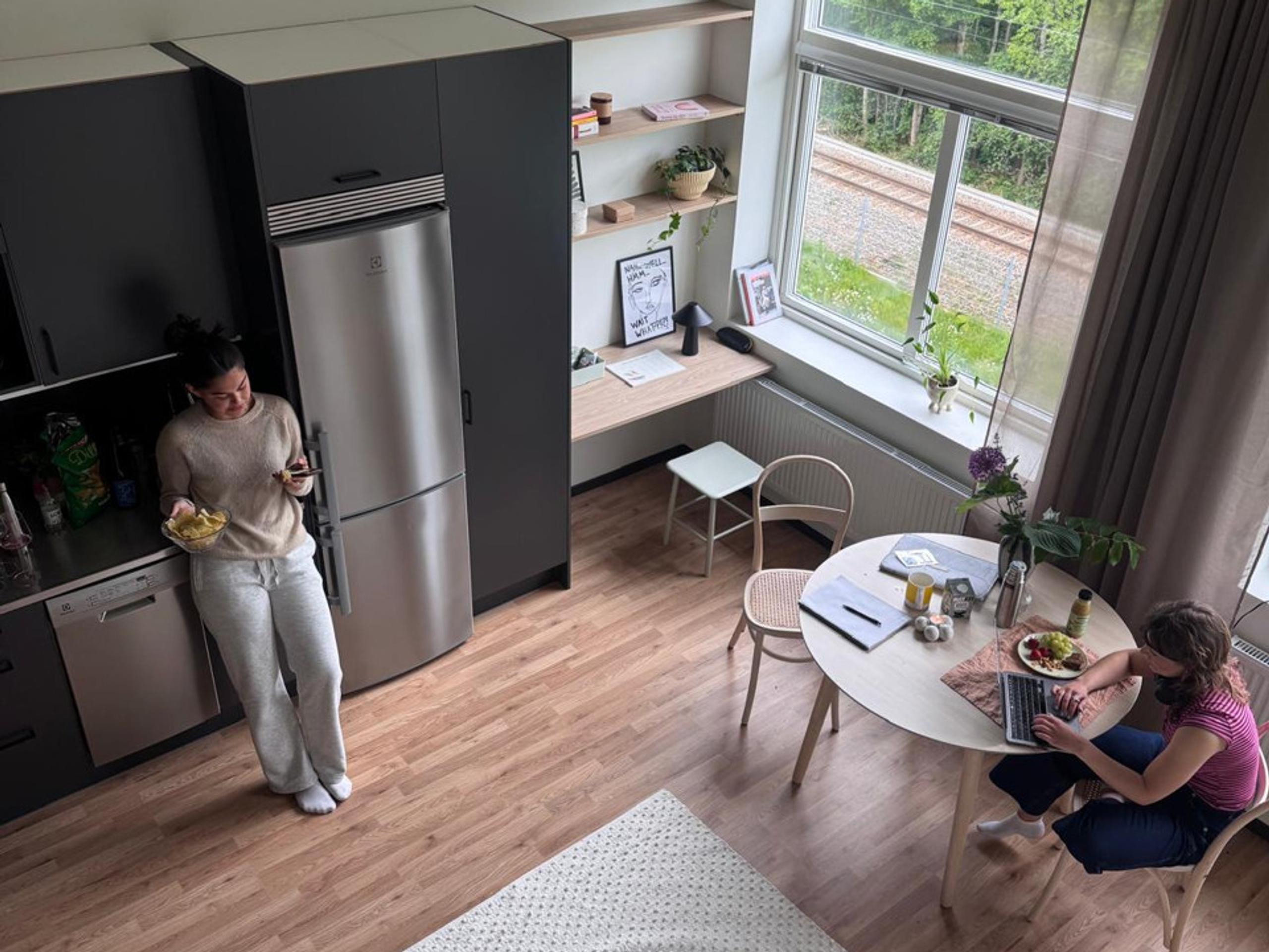 Two young women spending time together in a student apartment. One is standing in the kitchen and the other is sitting at the computer