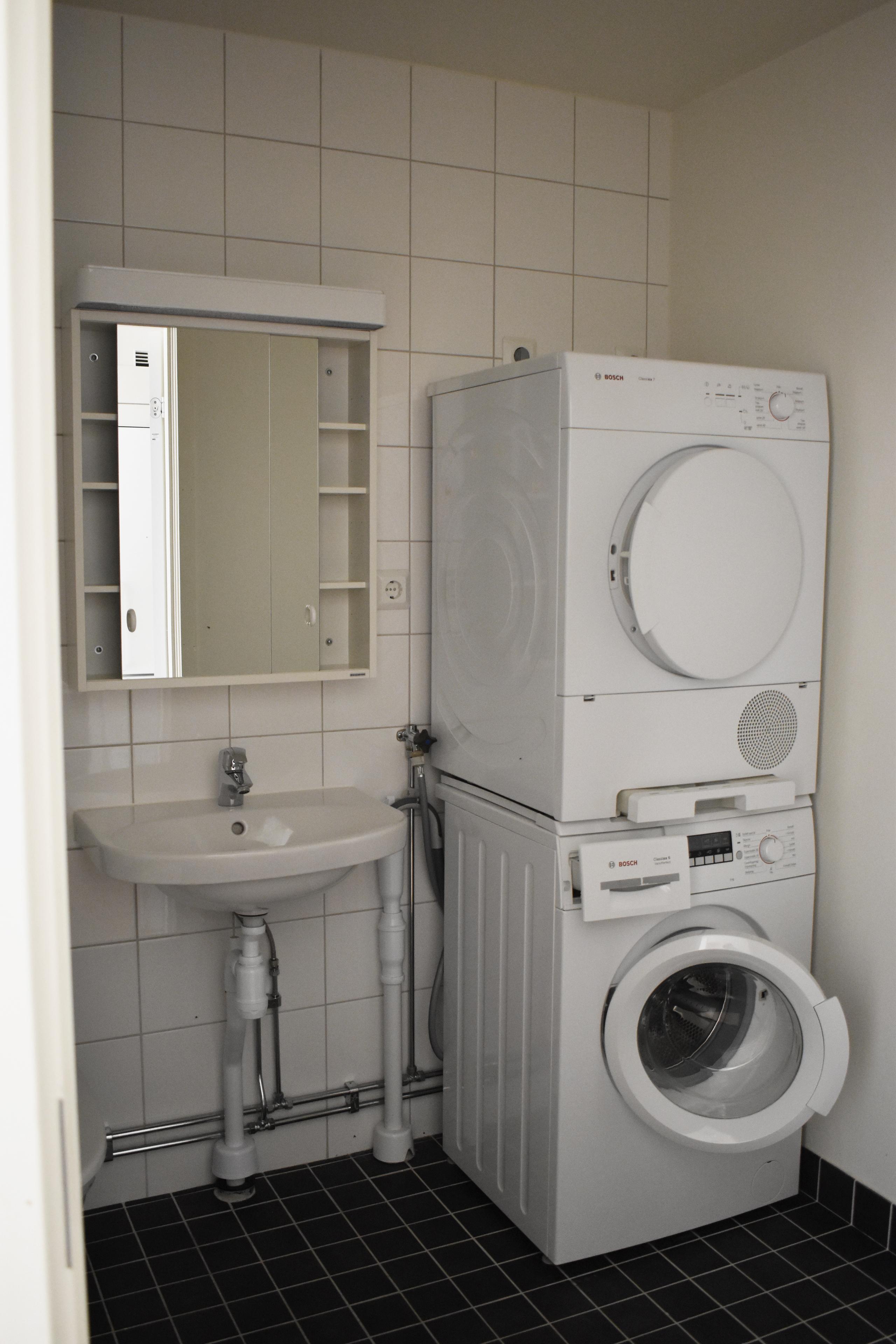 Student apartment. Fully tiled bathroom with washbasin, bathroom cabinet and a washing machine with a tumble dryer on top.