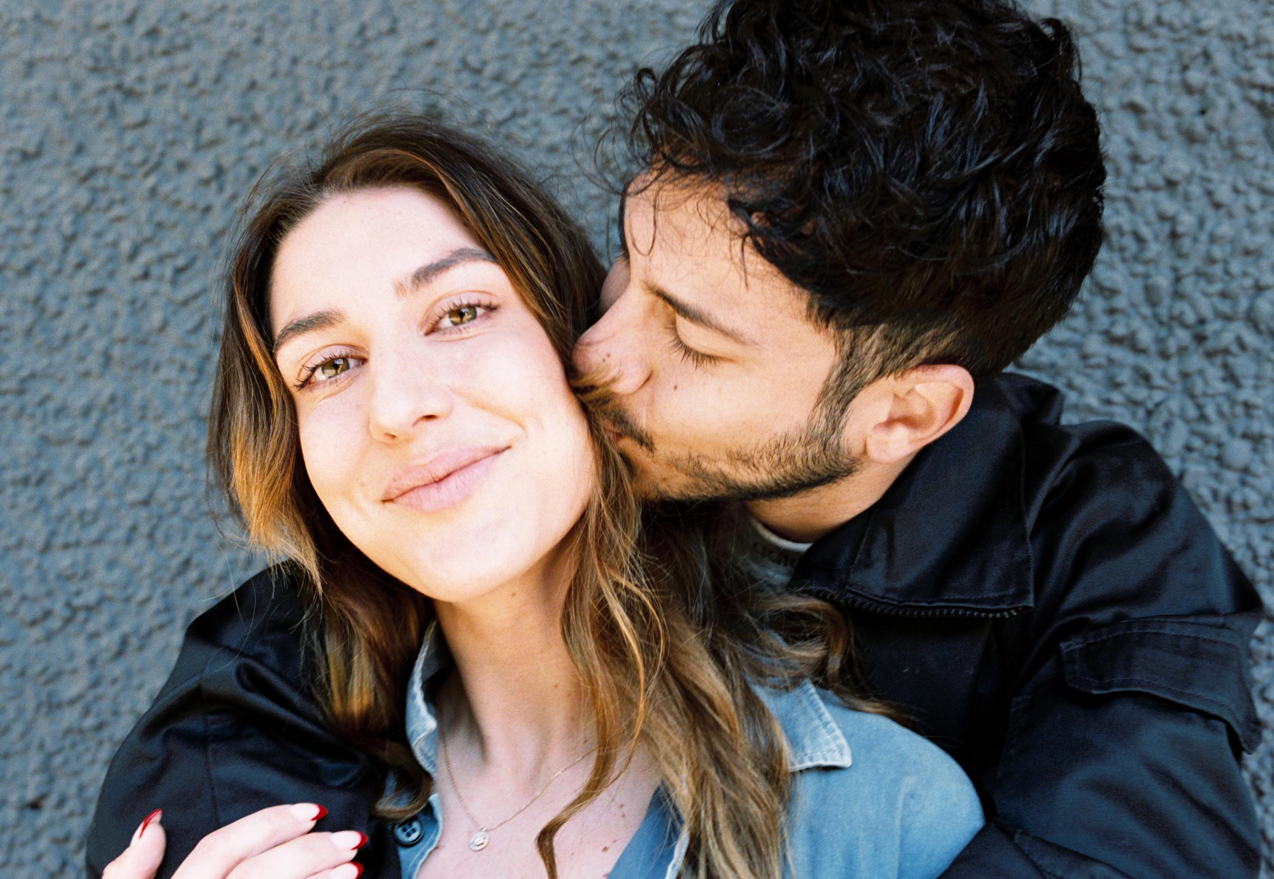 A young man stands behind a young woman and kisses her on the cheek.