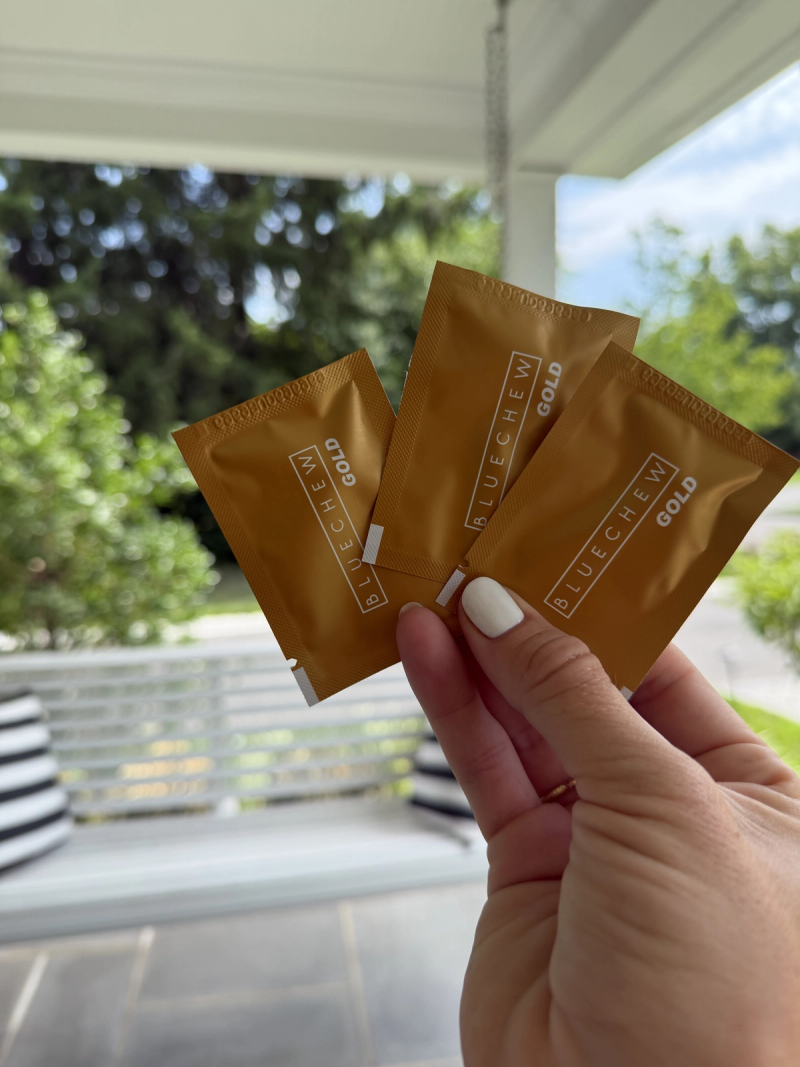 womens hand holding three bluechew gold packets on porch with greenery in background