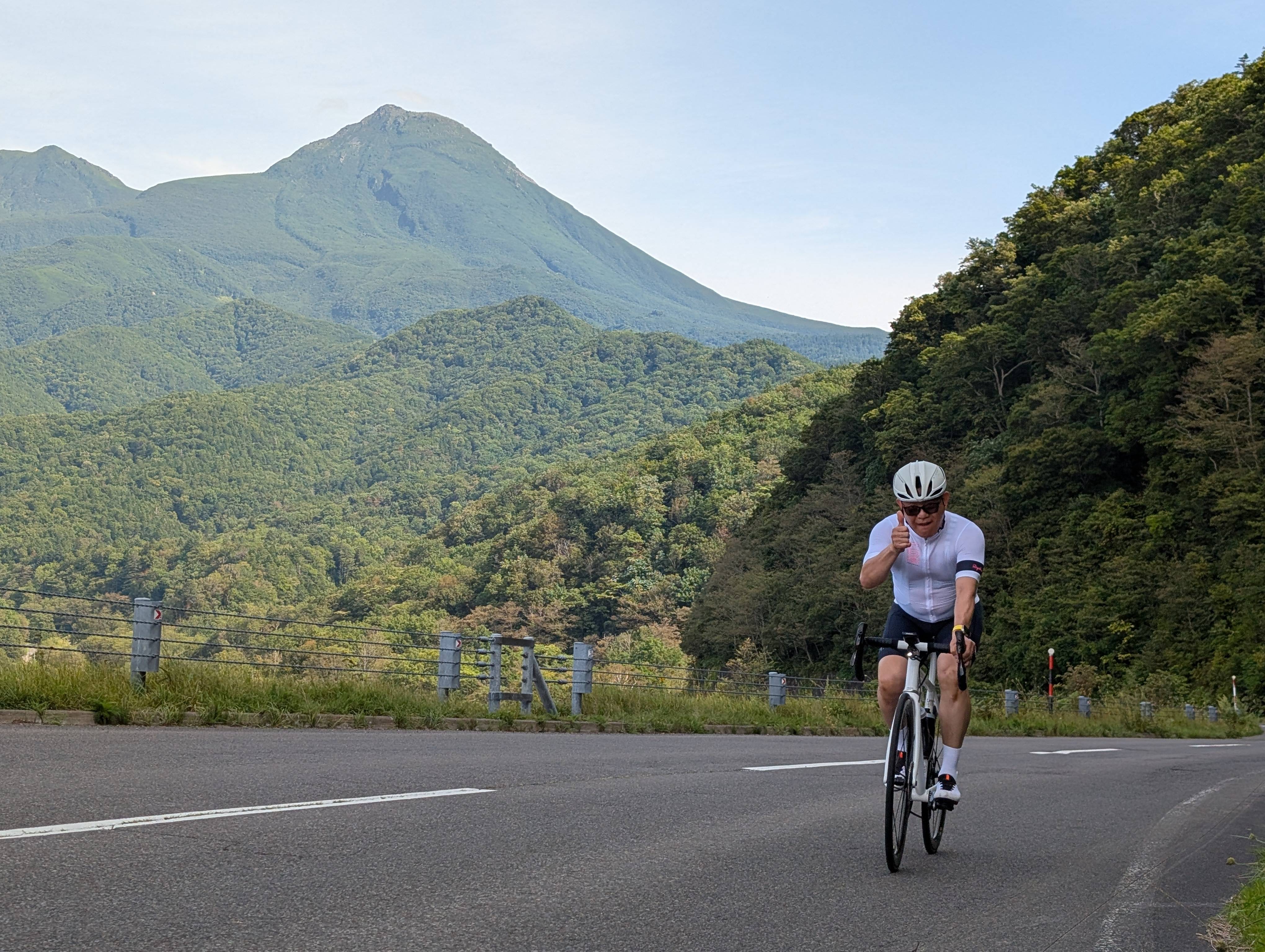 A man cycles up a mountain pass, giving the camera a thumbs-up. It is a lovely sunny day and a mountain rises in the distance behind him.