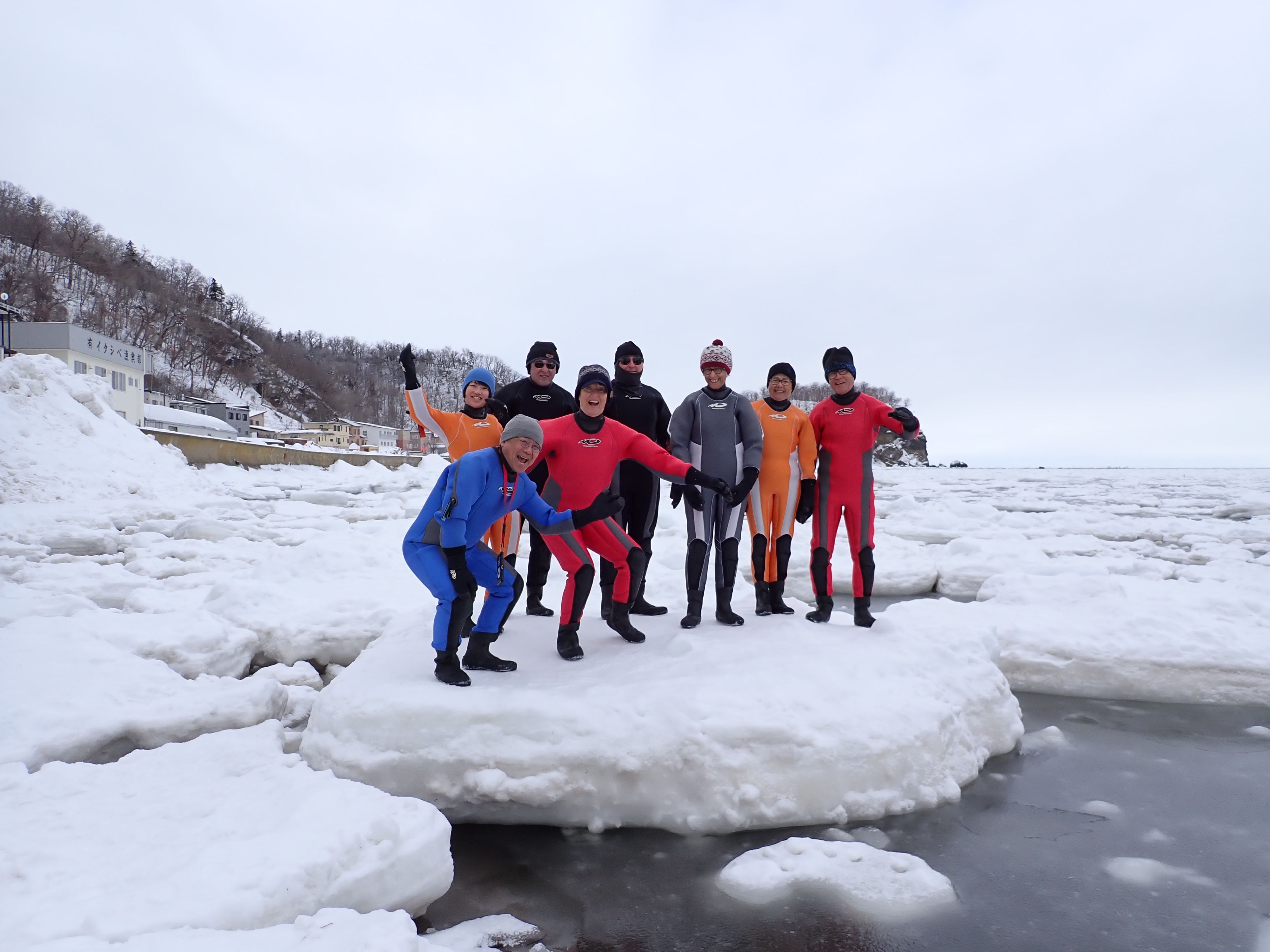 A group of people in drysuits smile and pose at the camera. They are standing on a floe of drift ice, which is floating in the ocean. Several more floes are visible behind them as far as the eye can see.