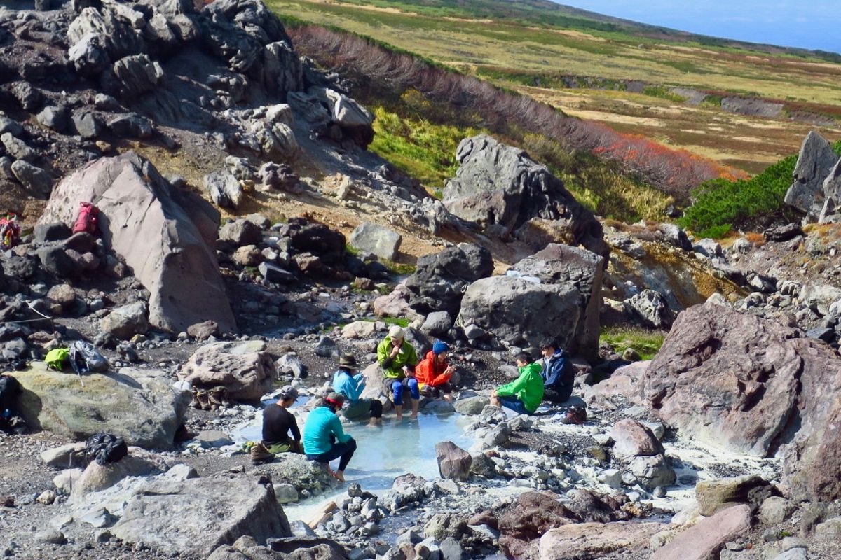 A group of hikers takes a break at Nakadake Onsen in Daisetsuzan, soaking their feet in a wild, milky-blue hot spring pool surrounded by large volcanic rocks and alpine tundra.