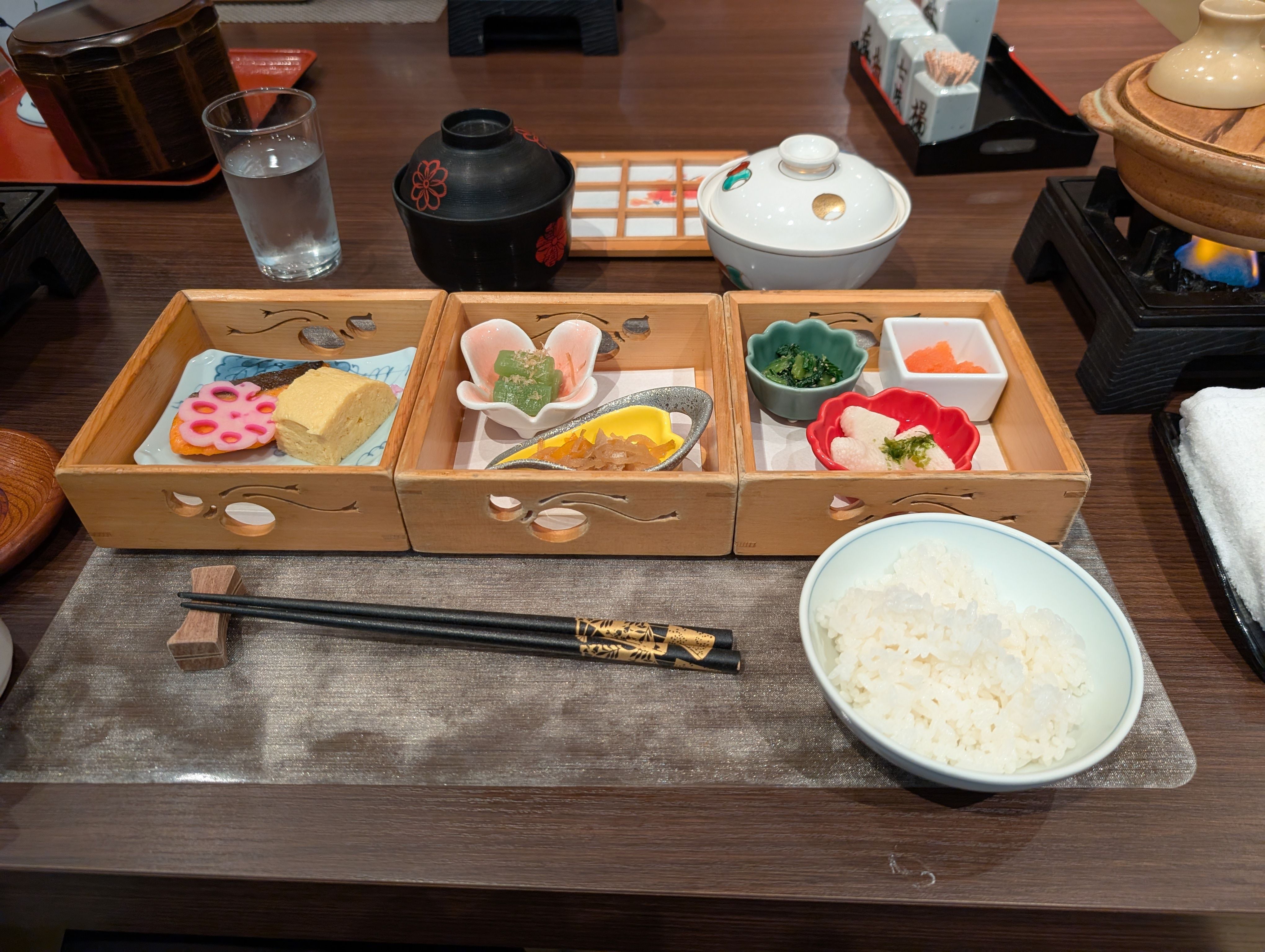 A traditional ryokan dinner is served in three wooden boxes on a dark table. The Japanese meal includes grilled salmon, tamagoyaki, pickles, and small side dishes in colourful ceramic bowls, accompanied by white rice, miso soup, and green tea.