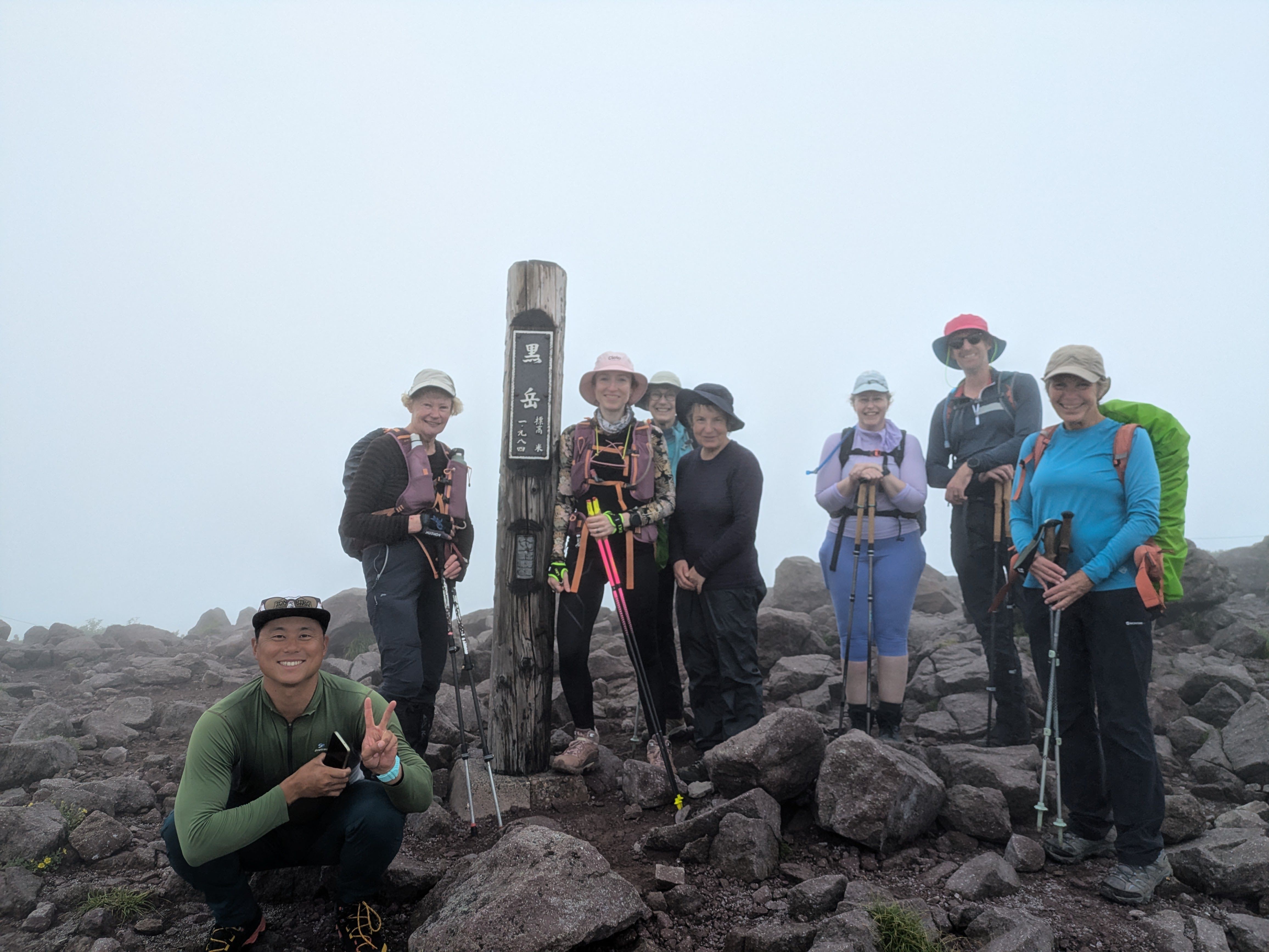 A group of hikers stand next to a pole at the summit of Mt. Kurodake in Hokkaido. The pole reads, in Japanese, "Mt. Kurodake, 1980m".