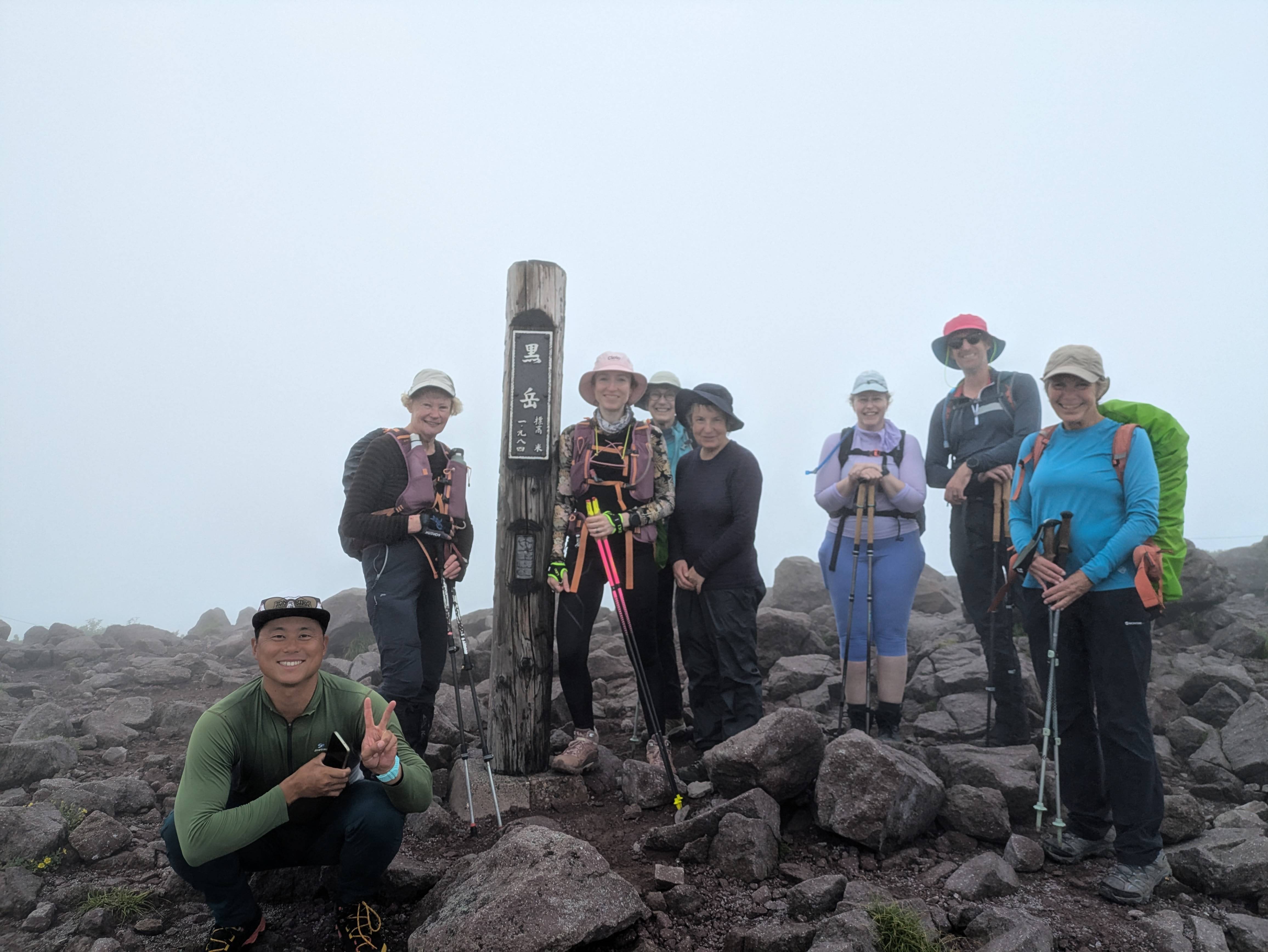 A group of hikers stand next to a pole at the summit of Mt. Kurodake in Hokkaido. The pole reads, in Japanese, "Mt. Kurodake, 1980m".