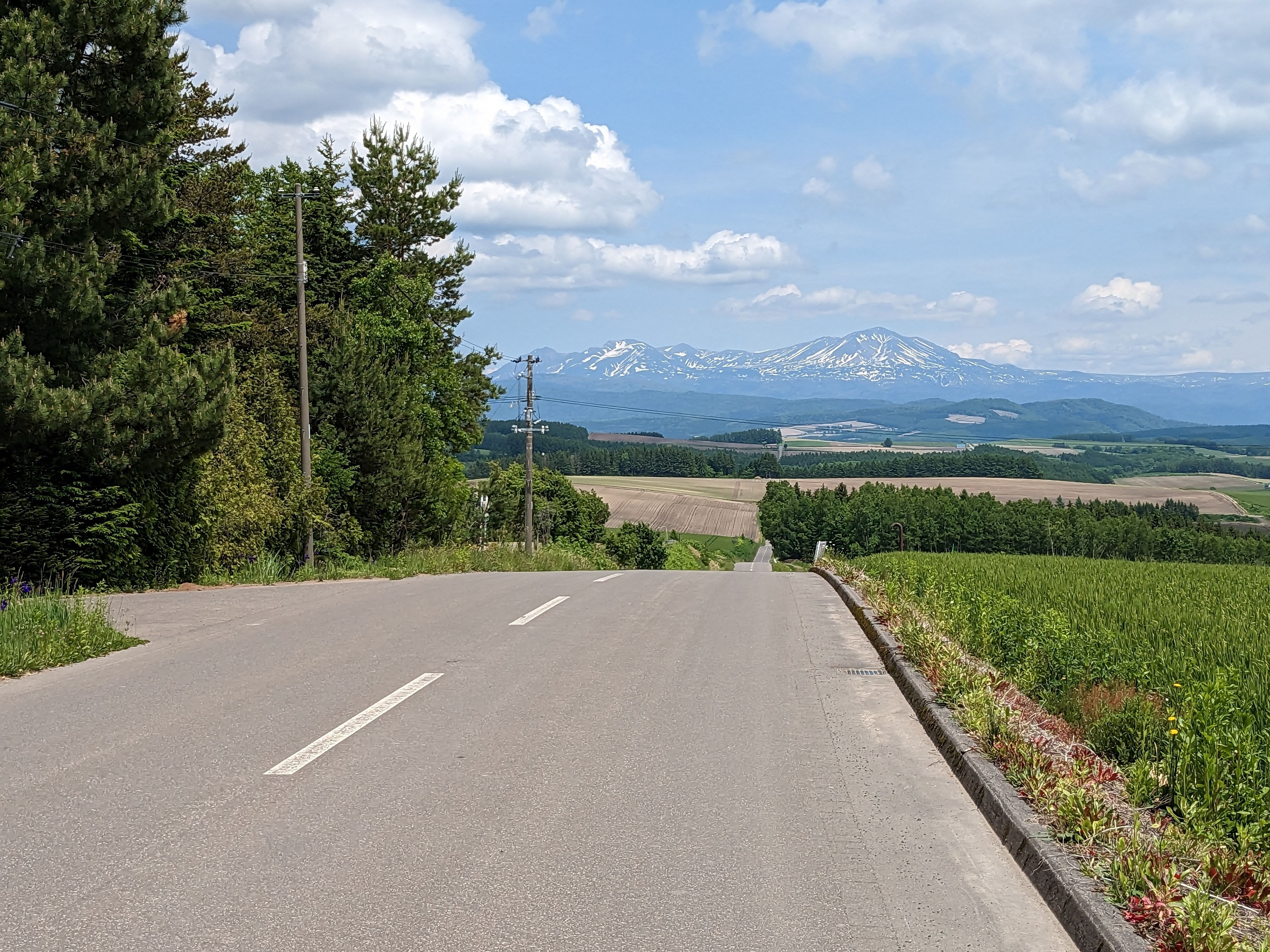 A view of Mt. Asahidake, the tallest mountain in Japan, in the distance. In the foreground, the crest of a hilly road rises towards the viewer.