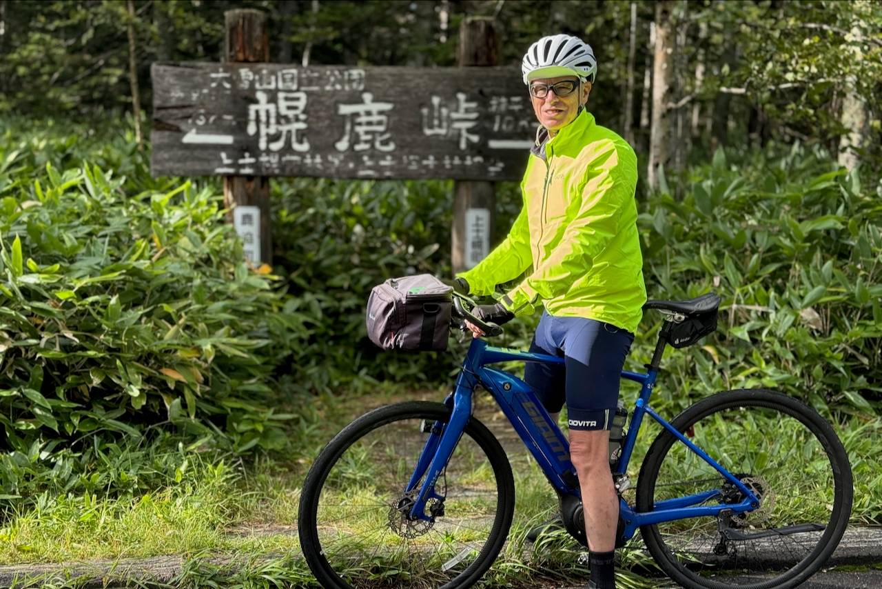 A male guest on an Adventure Hokkaido bike tour poses with his e-bike at Horoka Pass in Daisetsuzan National Park, Japan. He is wearing a helmet and a bright yellow jacket.