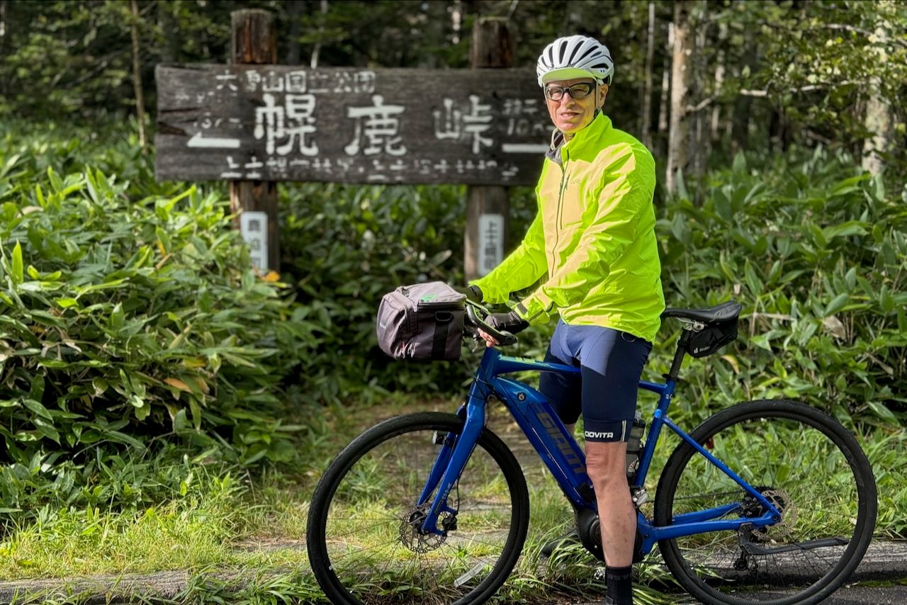 A male guest on an Adventure Hokkaido bike tour poses with his e-bike at Horoka Pass in Daisetsuzan National Park, Japan. He is wearing a helmet and a bright yellow jacket.