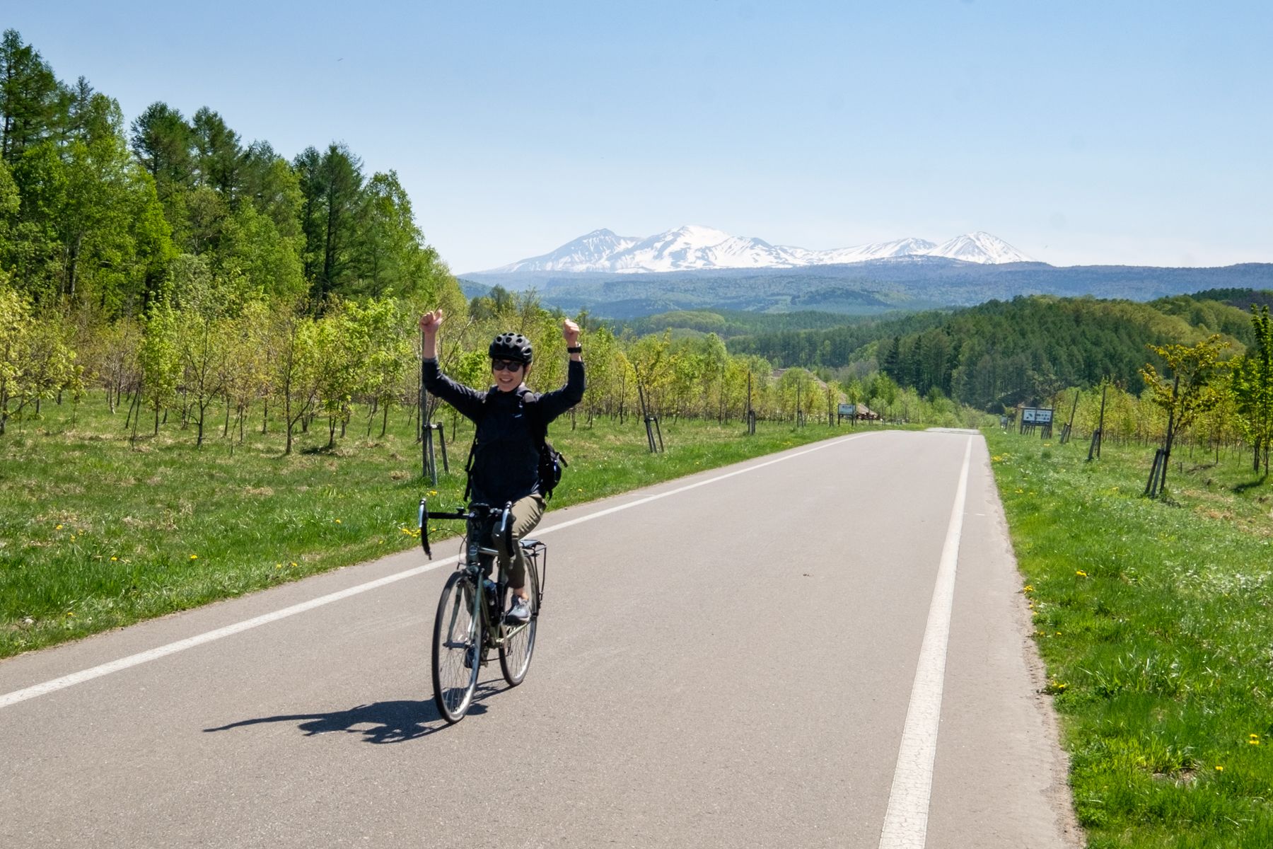 A cyclist celebrates with arms raised while riding along a paved road in Hokkaido. Lush green forests line the path, leading towards the dramatic, snow-capped peaks of the Daisetsuzan mountain range under a clear blue sky.