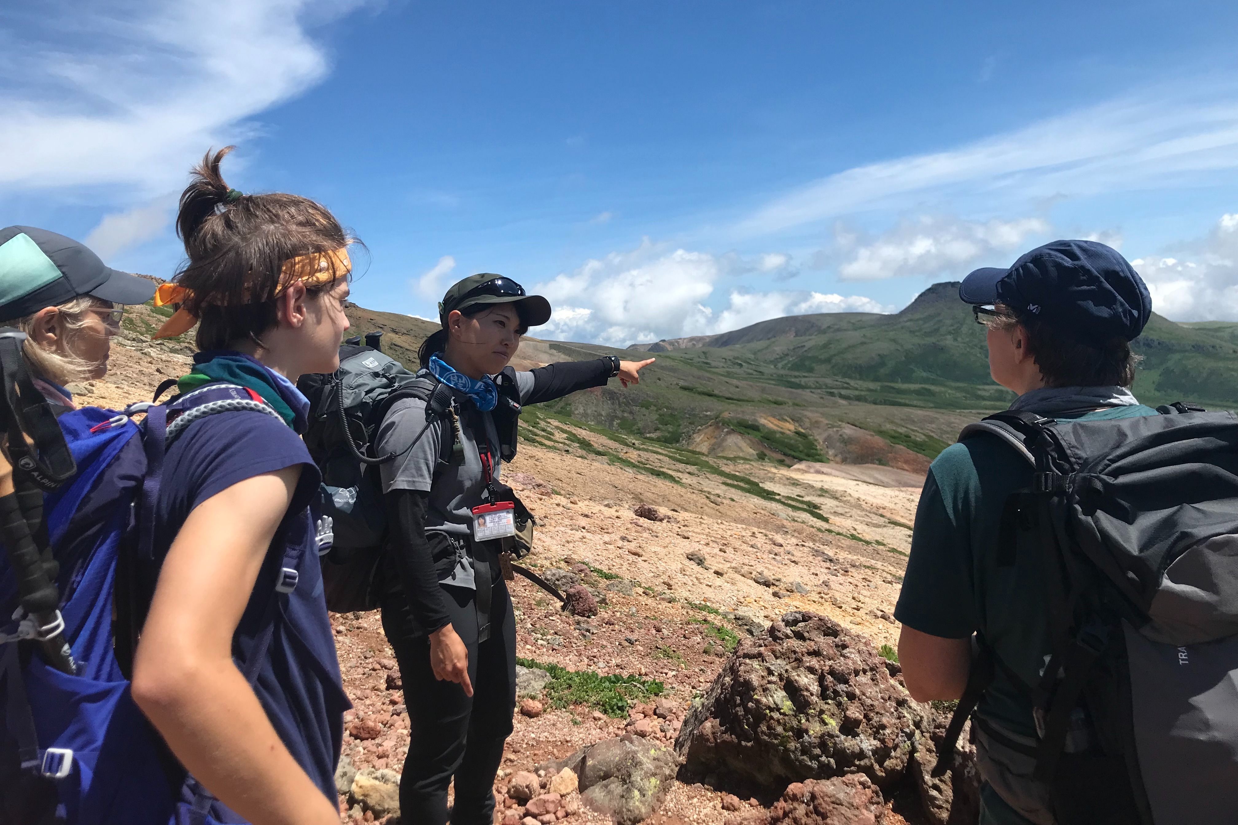 A hiking guide points towards a mountain while she explains the Daisetsuzan landscape to guests. They are stood on a rocky slope with expansive views under a blue sky.