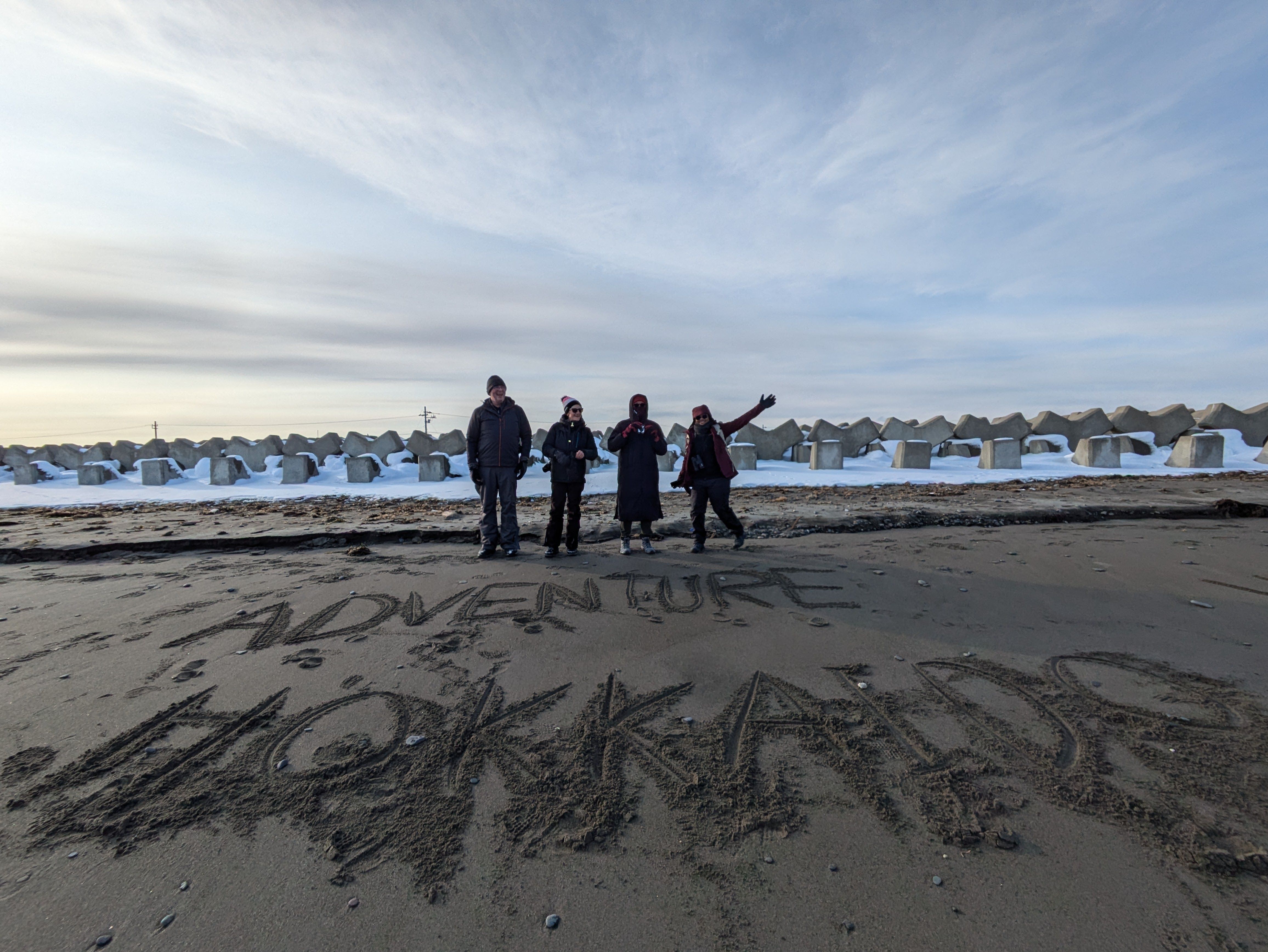 A group of four people pose and wave behind writing on a snowy winter beach. In the sand, they have written the words "ADVENTURE HOKKAIDO".