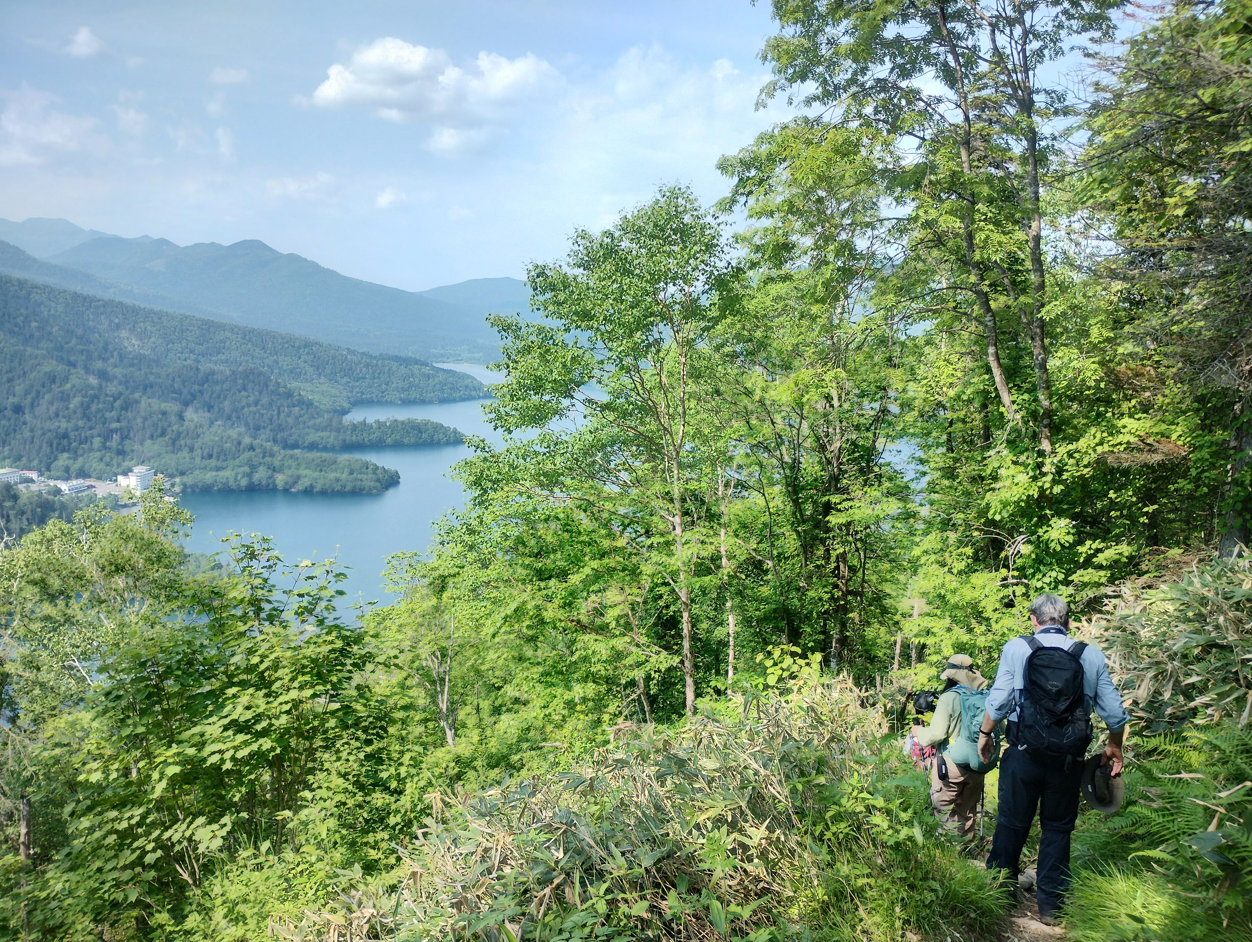 A group of hikers descend a mountain trail on a sunny day. In the distance, Lake Shikaribetsu is visible.