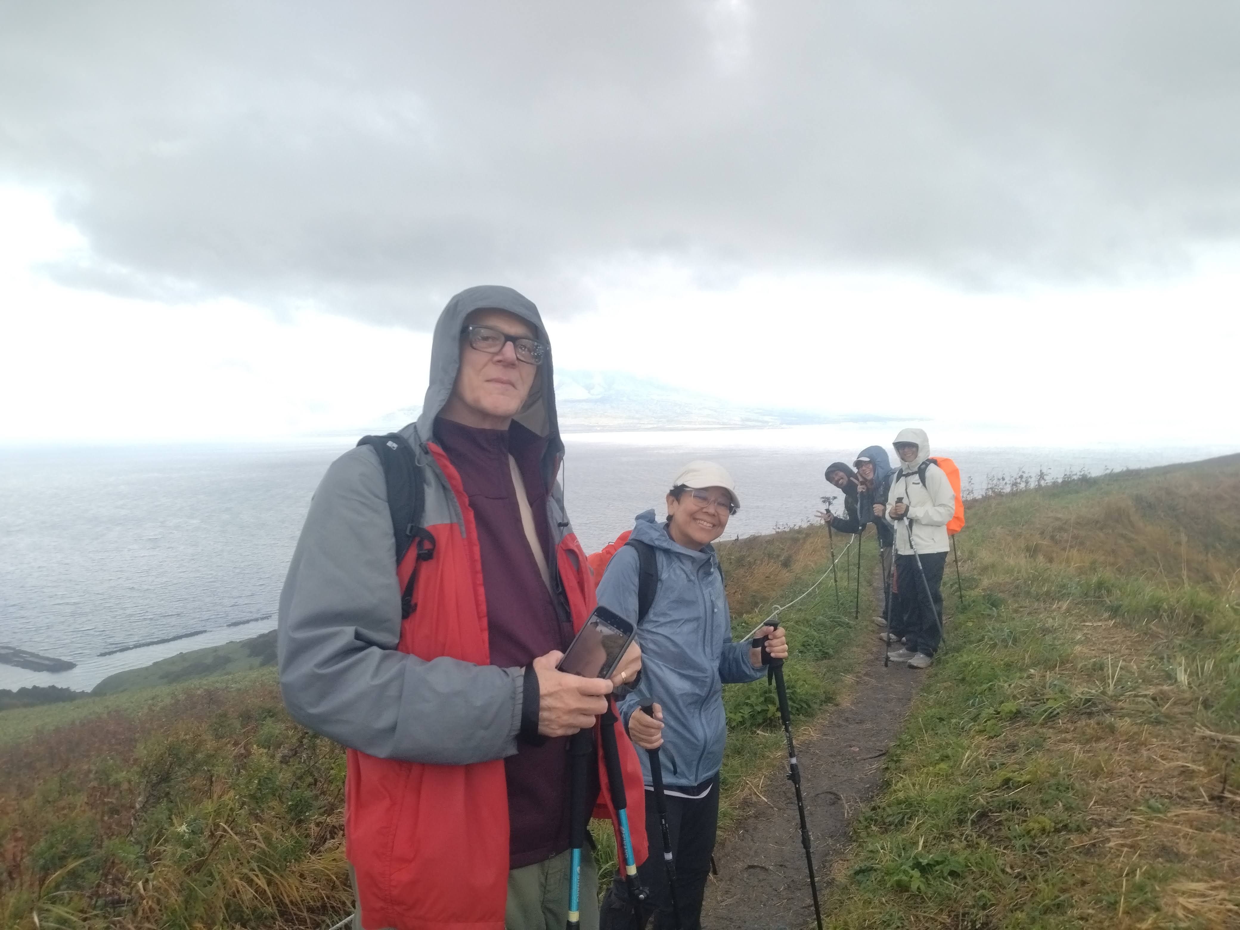Five smiling hikers in colourful rain gear and holding trekking poles pause along the narrow dirt path of the Momoiwa Trail on Rebun Island. They are standing on a steep, grassy coastal hillside overlooking the ocean, with a distant misty coastline visible under a cloudy sky.